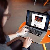 Eine Person mit langen, dunklen Haaren sitzt an einem runden, orangefarbenen Tisch und arbeitet an einem Laptop. Auf dem Bildschirm ist der Newsletter der Stiftung Zollverein zu sehen, mit einem Bild der beleuchteten Rolltreppe auf dem Welterbe Zollverein und dem Text: Staunen erlaubt: ein einzigartiger Ort für Erlebnisse. Neben dem Laptop stehen eine Tasse Cappuccino mit Löffel und ein Smartphone auf dem Tisch. Die Szene wirkt ruhig und konzentriert, in moderner Café-Atmosphäre.