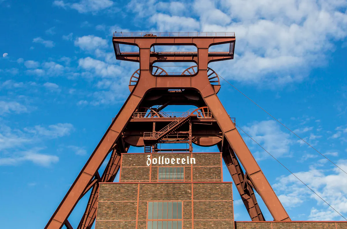 Das Bild zeigt das markante Doppelbock-Fördergerüst der Zeche Zollverein in Essen bei Tageslicht. Das massive, rostrote Stahlgerüst ragt in den blauen Himmel mit vereinzelten weißen Wolken. Darunter ist das ehemalige Maschinenhaus aus dunklem Backstein zu sehen, mit dem weißen Schriftzug: Zollverein. Das Fördergerüst ist ein bekanntes Wahrzeichen des UNESCO-Welterbes.