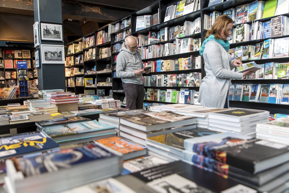 Innenaufnahme des Museumsshops mit zwei Personen, die in Büchern stöbern. Im Vordergrund sind mehrere Tische mit aufgestapelten Büchern zu sehen, viele davon mit kulturellen, historischen oder regionalen Themen. Im Hintergrund befinden sich hohe, dunkle Bücherregale, die dicht mit Magazinen, Katalogen und weiteren Druckerzeugnissen gefüllt sind. Die Atmosphäre ist ruhig und einladend.