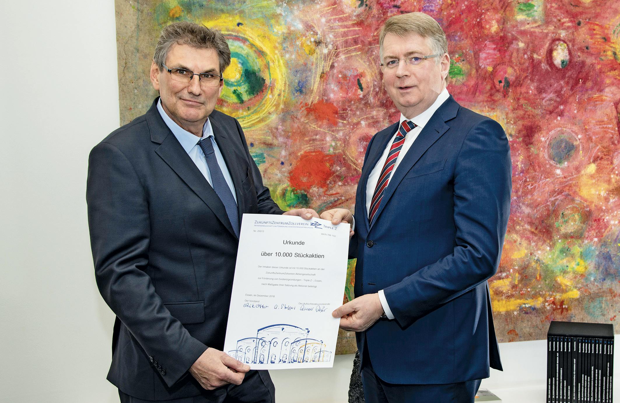 Zwei Männer in Anzügen stehen nebeneinander vor einem farbenfrohen, abstrakten Gemälde und halten gemeinsam ein großes Zertifikat. Beide schauen freundlich in die Kamera. Der Mann links trägt einen dunkelgrauen Anzug mit blauer Krawatte und Brille, der Mann rechts einen dunkelblauen Anzug mit rot-blau gestreifter Krawatte. Die Urkunde, die sie in den Händen halten, stammt vom Zukunftszentrum Zollverein und bestätigt die Übergabe von 10.000 Stückaktien. Im Vordergrund unten ist eine gezeichnete Illustration eines historischen Gebäudes zu sehen, rechts im Bild steht ein Regal mit schwarzen Buchrücken. Die Szene wirkt feierlich und offiziell.