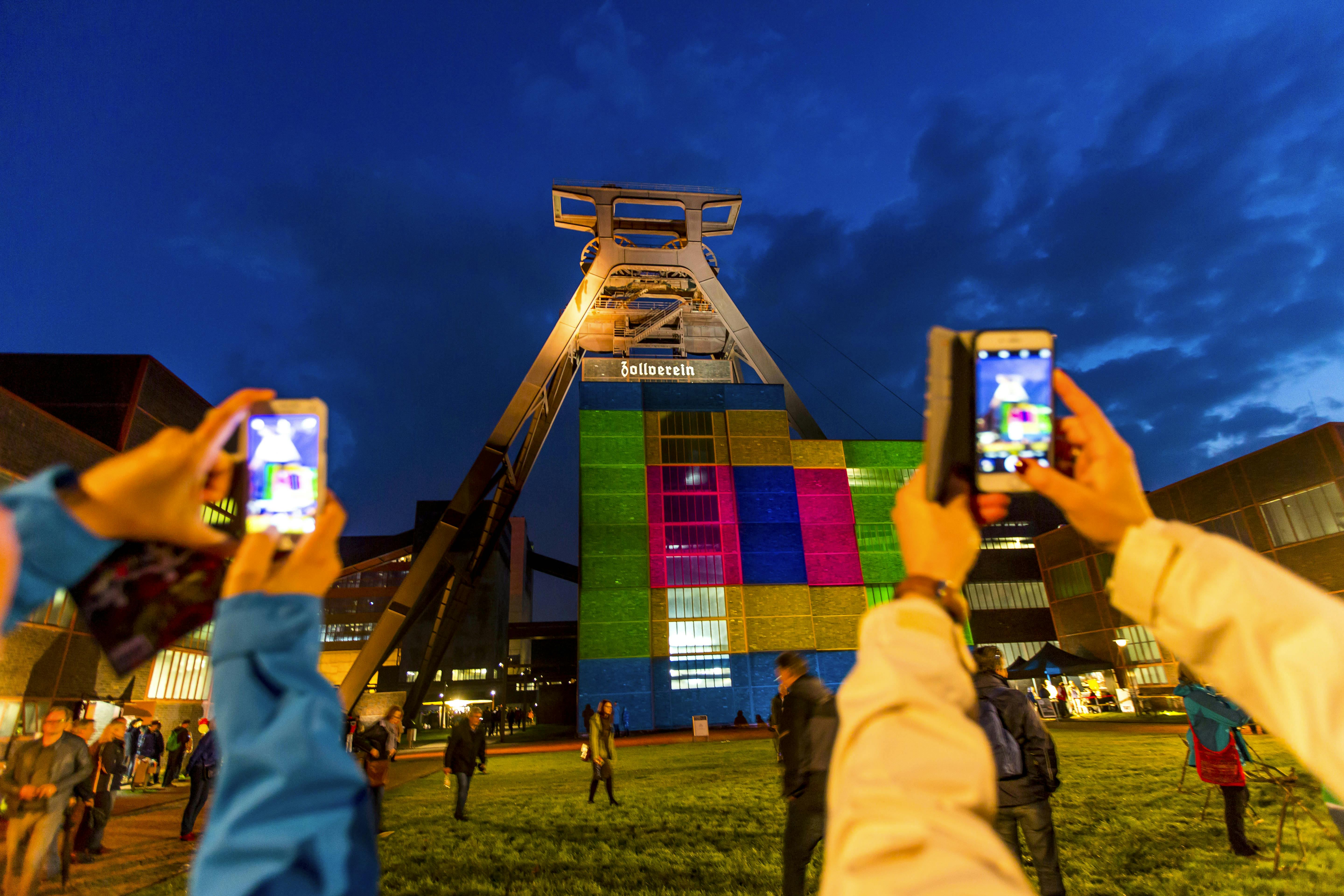 Abendaufnahme auf dem UNESCO-Welterbe Zollverein. Im Mittelpunkt steht das beleuchtete Fördergerüst von Schacht XII. Die Fassade des dahinterliegenden Gebäudes ist bunt illuminiert mit quadratischen Farbflächen in Grün, Blau, Pink und Gelb. Im Vordergrund halten zwei Personen Smartphones hoch und fotografieren das Lichtspektakel. Der Himmel ist dunkelblau, erste Sterne sind sichtbar. Weitere Menschen schlendern über die Rasenfläche oder stehen in kleinen Gruppen. Die Szene vermittelt eine lebendige Atmosphäre bei einem abendlichen Event.