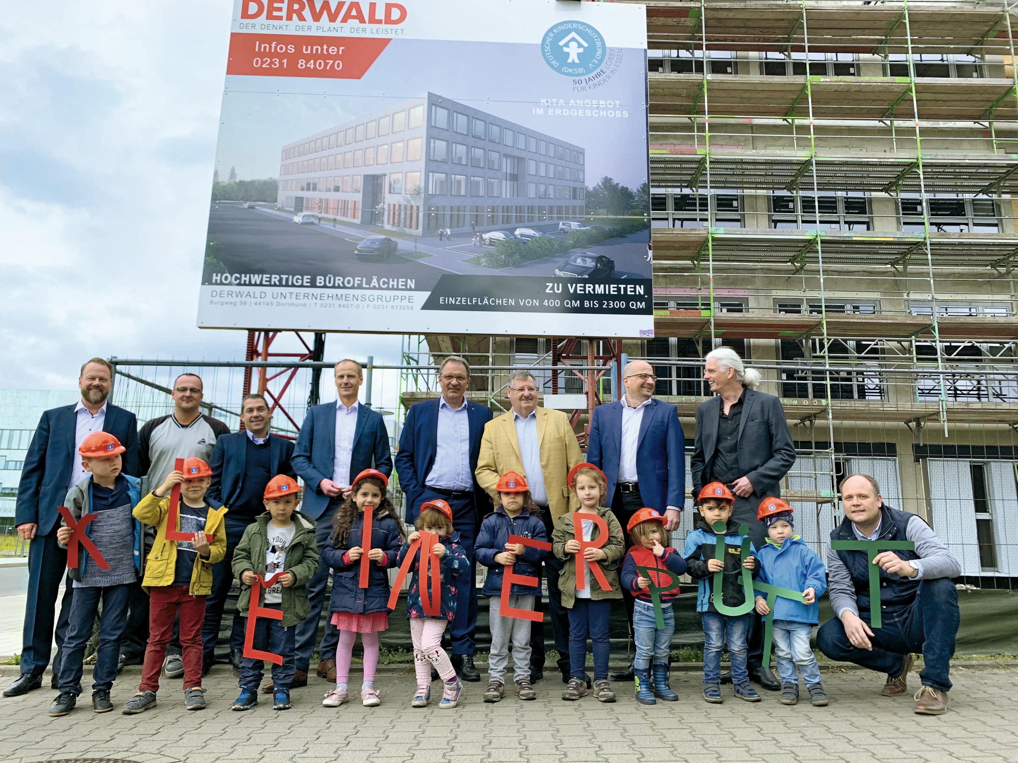 Vor einer Baustelle mit einem eingerüsteten Neubau stehen mehrere Männer in Anzügen sowie eine Gruppe von Kindern mit roten Bauhelmen. Die Kinder halten große, bunte Buchstaben in den Händen, die gemeinsam den Namen KLEINER PÜTT ergeben. Im Hintergrund ist eine große Bautafel zu sehen: Sie wirbt für hochwertige Büroflächen und zeigt das geplante Gebäude mit dem Hinweis auf ein Kita-Angebot im Erdgeschoss. Die Szene zeigt den symbolischen Baustart mit Beteiligung von Kindern und Projektverantwortlichen.