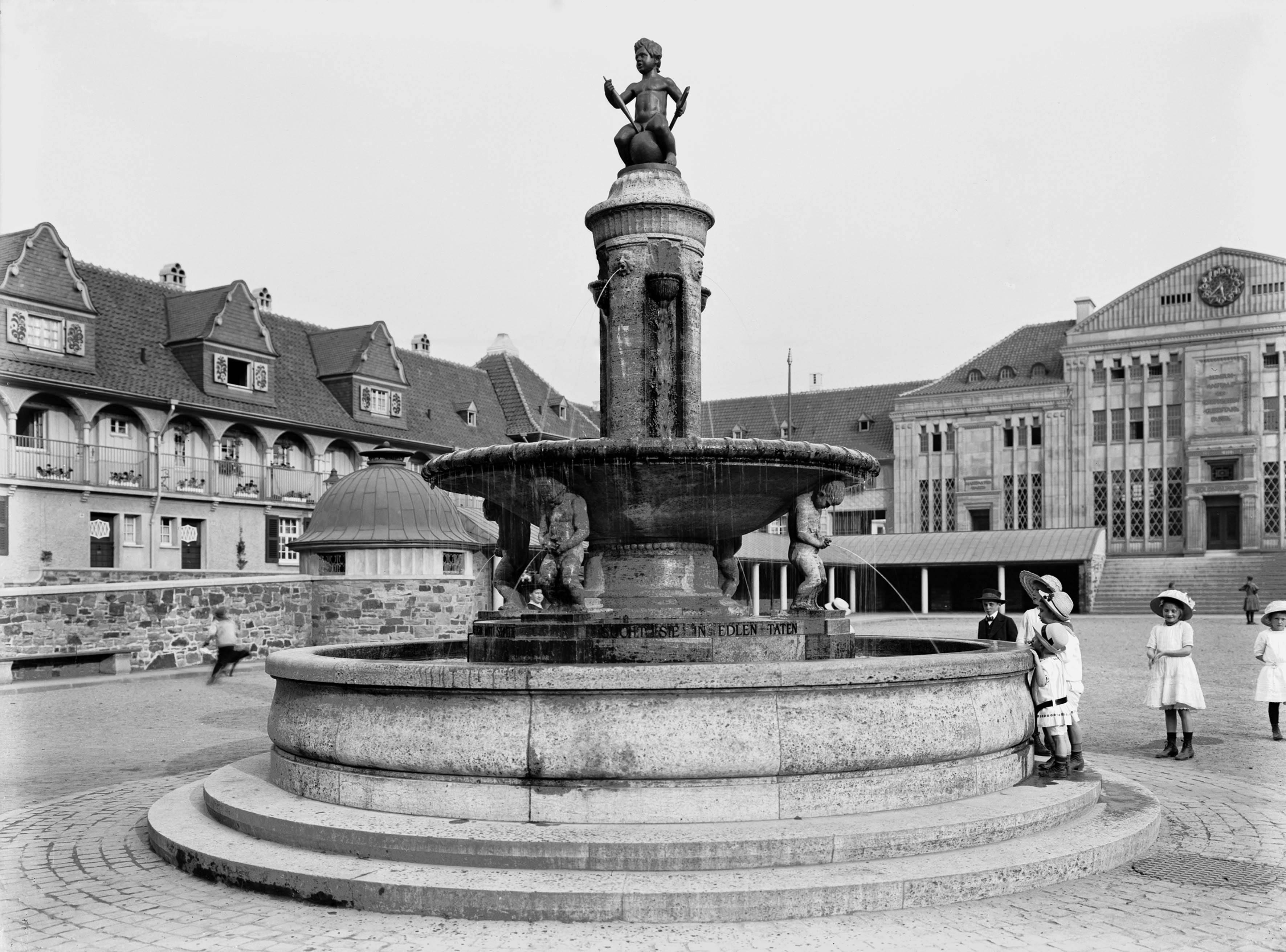 Historische Schwarzweißaufnahme des Brunnens auf dem zentralen Marktplatz der Margarethenhöhe in Essen. Der Brunnen ist mit mehreren Wasserspeiern und einer Skulptur eines Kindes auf der Spitze verziert. Im Hintergrund sind Gebäude im reformerischen Heimatstil mit Rundbögen und Giebeldächern zu sehen. Vor dem Brunnen stehen mehrere Kinder in Kleidung aus der Zeit um 1915.