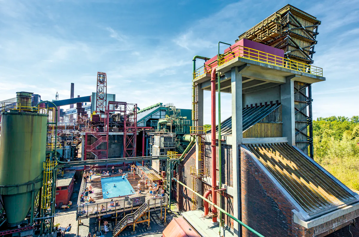 Das Bild zeigt das Gelände eines ehemaligen Industriekomplexes mit beeindruckender Industriearchitektur aus Stahl, Beton und Backstein. Im Zentrum befindet sich ein rechteckiges Schwimmbecken mit Holzdeck, auf dem zahlreiche Menschen bei sommerlichem Wetter entspannen oder baden. Umgeben ist das Becken von mehrstöckigen, teils rostigen Industrieanlagen, Rohren, Silos und Förderanlagen. Rechts im Bild ragt ein modernisiertes Gebäude mit hölzern verkleideten Elementen und gelbem Geländer empor. Im Hintergrund sind grüne Bäume und blauer Himmel mit leichten Wolken zu sehen. Das Bild vermittelt den Kontrast zwischen industrieller Vergangenheit und heutiger Freizeitnutzung.