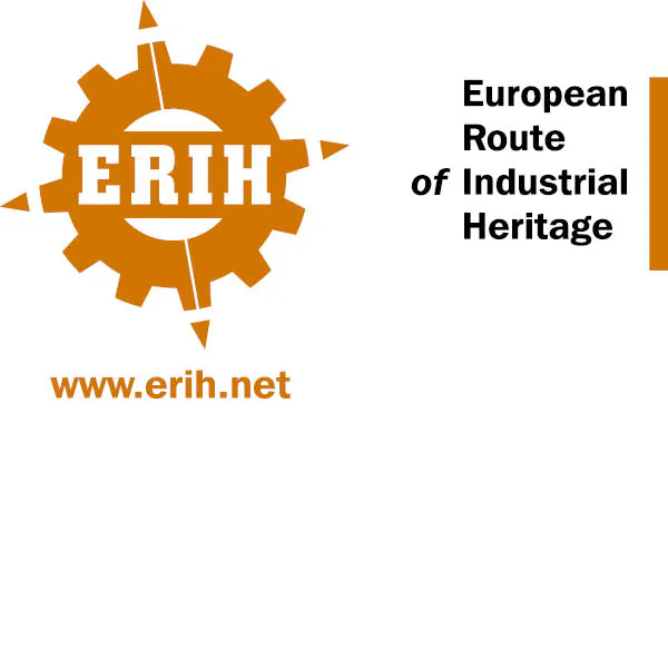 Das Bild zeigt das Logo der ERIH – European Route of Industrial Heritage (Europäische Route der Industriekultur). Links ist ein stilisiertes Zahnrad in Orange abgebildet, in dessen Mitte in weißen Großbuchstaben steht: ERIH. Das Zahnrad hat acht Zähne und erinnert an industrielle Technik. Darunter befindet sich die orangefarbene Internetadresse: www.erih.net. Rechts daneben steht in schwarzer Schrift: European Route of Industrial Heritage. Das Design vermittelt den industriellen Charakter des Netzwerks, das bedeutende Orte der Industriekultur in Europa verbindet.