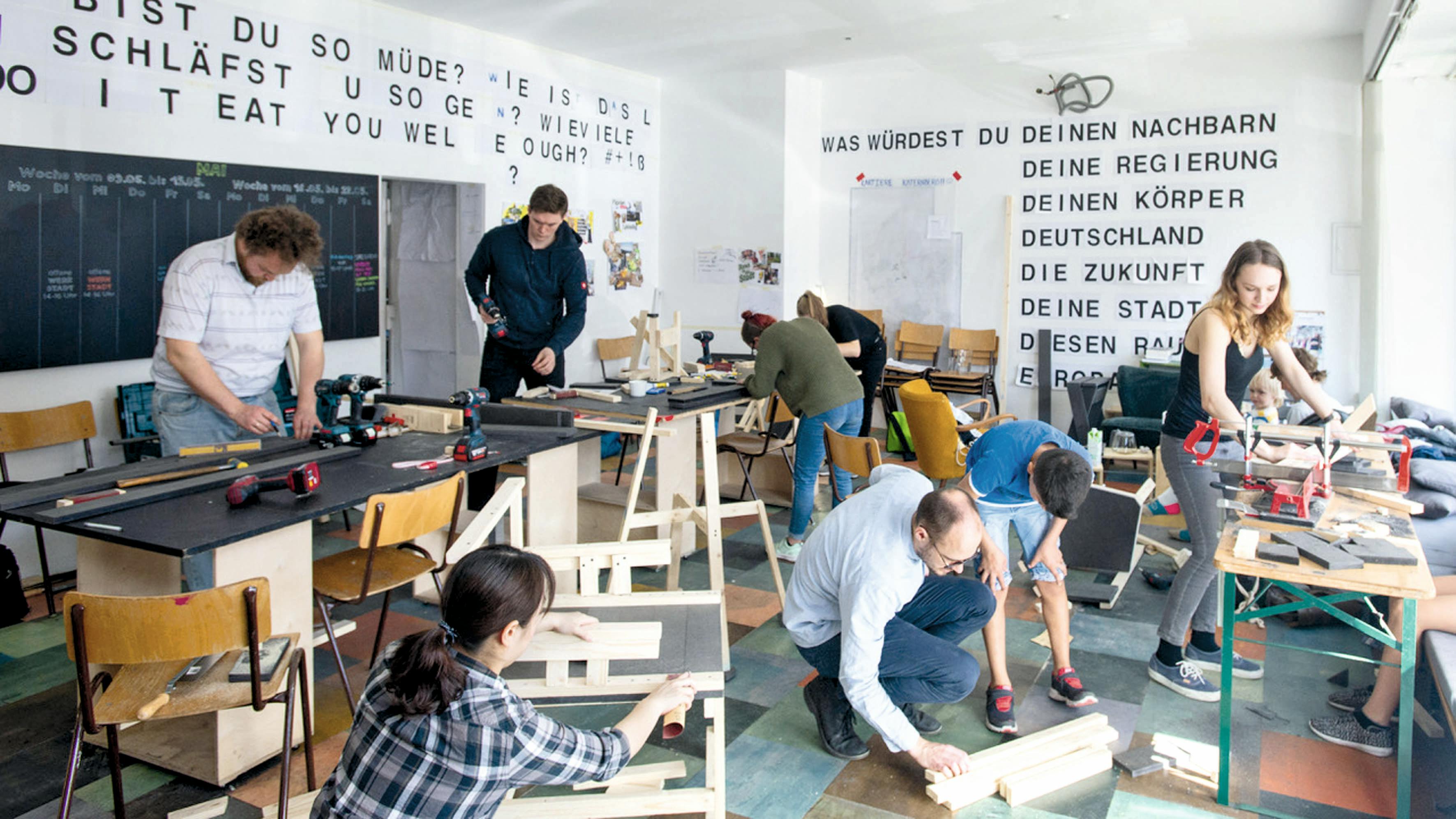 In einem hellen Raum mit großen Fenstern arbeiten mehrere Menschen konzentriert an verschiedenen Holzprojekten. Auf Tischen und Werkbänken liegen Werkzeuge wie Akkuschrauber, Zwingen und Holzlatten. Einige Personen stehen oder sitzen auf Stühlen, andere knien auf dem Boden, um Werkstücke zu bearbeiten. Im Hintergrund sind an der weißen Wand großformatige Fragen in schwarzer Schrift zu lesen, etwa: Was würdest du deinen Nachbarn, deine Regierung, deinen Körper, Deutschland, die Zukunft… – eine Anregung zur Reflexion. Die Atmosphäre ist lebendig und kreativ, der Raum wirkt wie eine offene Werkstatt oder ein partizipatives Kunstprojekt.