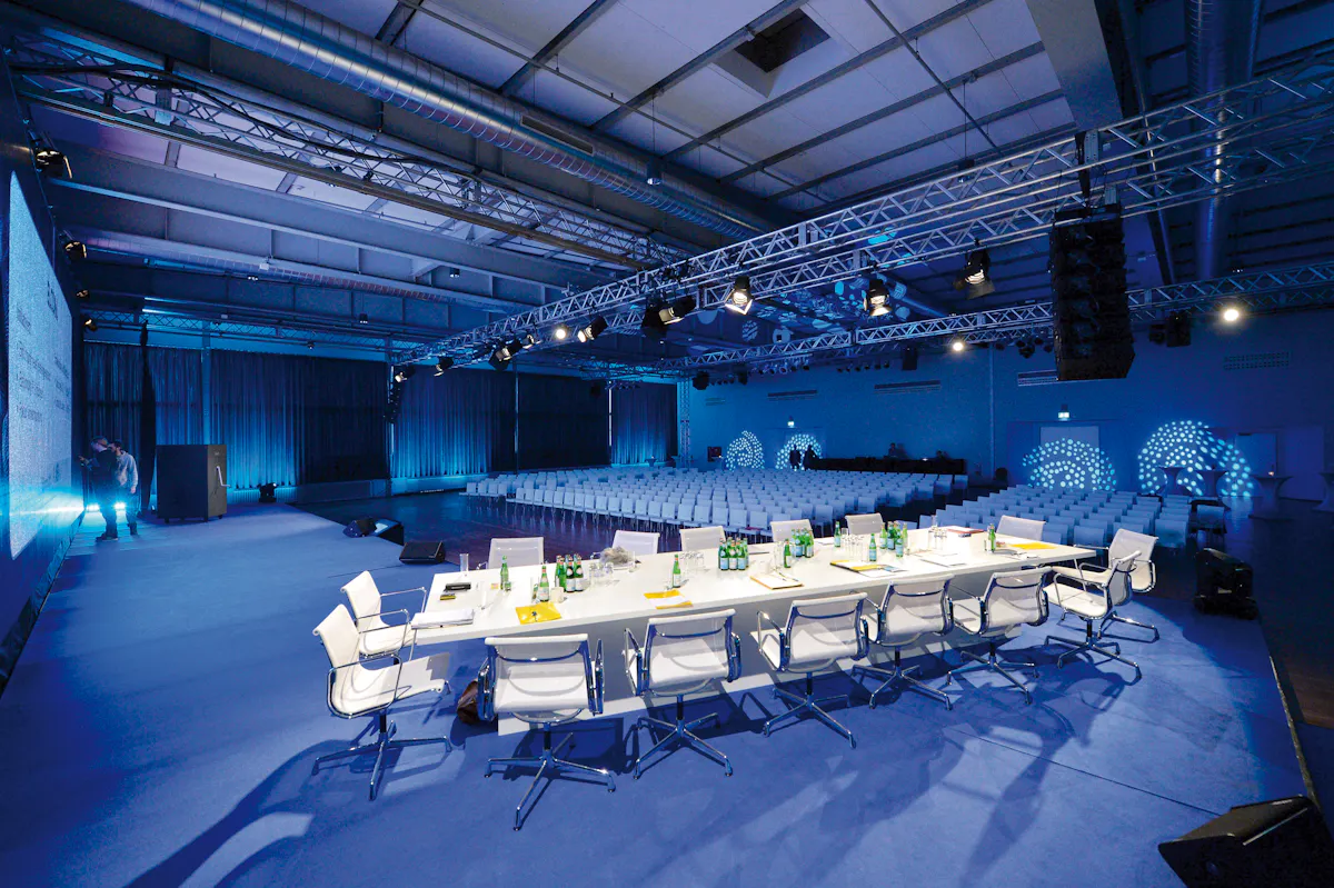 Das Bild zeigt einen großen, modern beleuchteten Konferenz- oder Veranstaltungssaal mit kühlem blauen Lichtambiente. Im Vordergrund steht ein weißer Konferenztisch, umgeben von weißen Designer-Drehstühlen. Auf dem Tisch stehen Wasserflaschen, Gläser, Notizblöcke und Namensschilder. Dahinter befinden sich viele gleichmäßig angeordnete weiße Stuhlreihen für das Publikum. Über dem Raum verläuft ein rigides Metallträger-System mit zahlreichen professionellen Scheinwerfern und Lautsprechern, die auf die Bühne und den Zuschauerbereich gerichtet sind. Links ist eine große Leinwand zu sehen, auf die Inhalte projiziert werden. Die rechte Wand ist mit dekorativen Lichtmustern aus kleinen Lichtpunkten beleuchtet. Die Atmosphäre wirkt professionell, hochwertig und technisch modern – geeignet für Konferenzen, Podiumsdiskussionen oder Preisverleihungen.