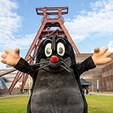 Vor dem markanten Förderturm der Zeche Zollverein in Essen steht eine große, freundliche Maulwurf-Figur mit ausgebreiteten Armen. Das Kostüm ist schwarz-grau mit weißem Gesicht, roten Wangen und einer roten Nase, die Figur hat große, lachende Augen und wirkt fröhlich. Der Hintergrund zeigt das historische Industriegelände bei blauem Himmel mit vereinzelten Wolken. Die Szene vermittelt einen spielerischen, einladenden Eindruck im Rahmen einer Veranstaltung für Kinder.