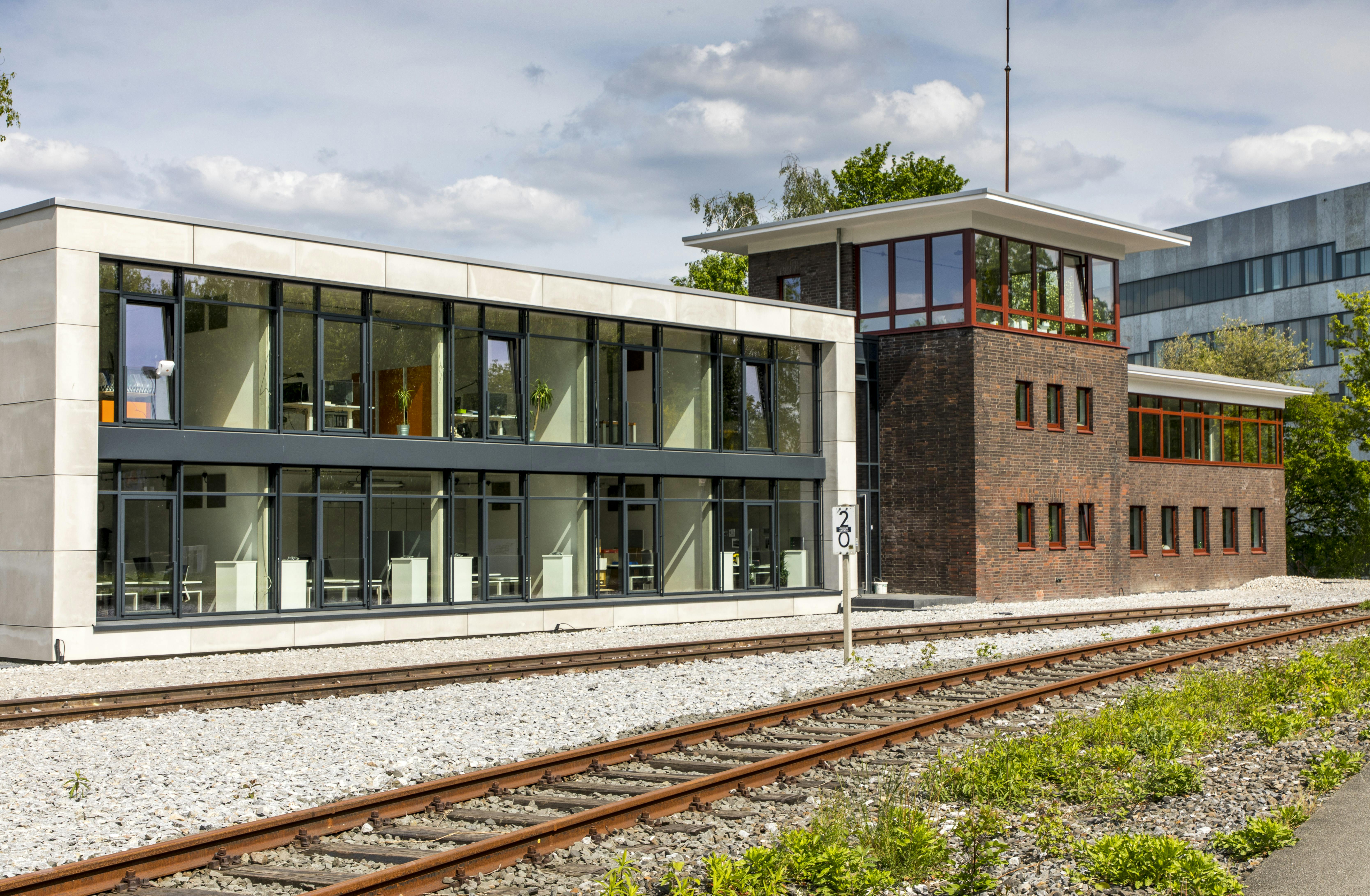 Das Bild zeigt ein modernes und ein historisches Gebäude auf dem Gelände des UNESCO-Welterbes Zollverein in Essen, direkt nebeneinander positioniert und parallel zu Eisenbahnschienen gelegen. Links im Bild befindet sich ein flacher, zweigeschossiger Neubau mit großen Glasfronten. Im Inneren sind helle, modern eingerichtete Schulungs- oder Konferenzräume mit weißen Tischen und Stühlen zu erkennen. Rechts daneben steht ein backsteinernes Stellwerksgebäude mit charakteristischen roten Fensterrahmen im Obergeschoss und einem auskragenden Flachdach. Die Gebäudekombination steht auf einem mit hellem Schotter bedeckten Boden neben alten Gleisen, was an die industrielle Nutzung des Areals erinnert. Im Hintergrund ist bei sonnigem Wetter grüne Vegetation und ein modernes Gebäude zu sehen.