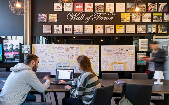 Das Bild zeigt einen modernen, kreativ gestalteten Besprechungsraum. Zwei Personen – ein Mann und eine Frau im gestreiften Pullover – sitzen an einem großen dunklen Konferenztisch mit schwarzen Stühlen. Sie unterhalten sich angeregt und schauen gemeinsam auf einen Laptop. Im Hintergrund ist eine Wand mit dem Schriftzug - Wall of Fame - zu sehen, über der zahlreich gerahmte Bilder, Urkunden und Plakate hängen. Darunter befinden sich zwei große, bunt illustrierte Mindmaps oder Präsentationstafeln. Rechts betritt ein Mann den Raum durch eine Tür, in der Hand hält er rote Ordner. Die Decke ist mit modernen, geometrischen Lampen geschmückt, die ein warmes Licht verbreiten. Der Raum wirkt kreativ, einladend und kommunikativ.