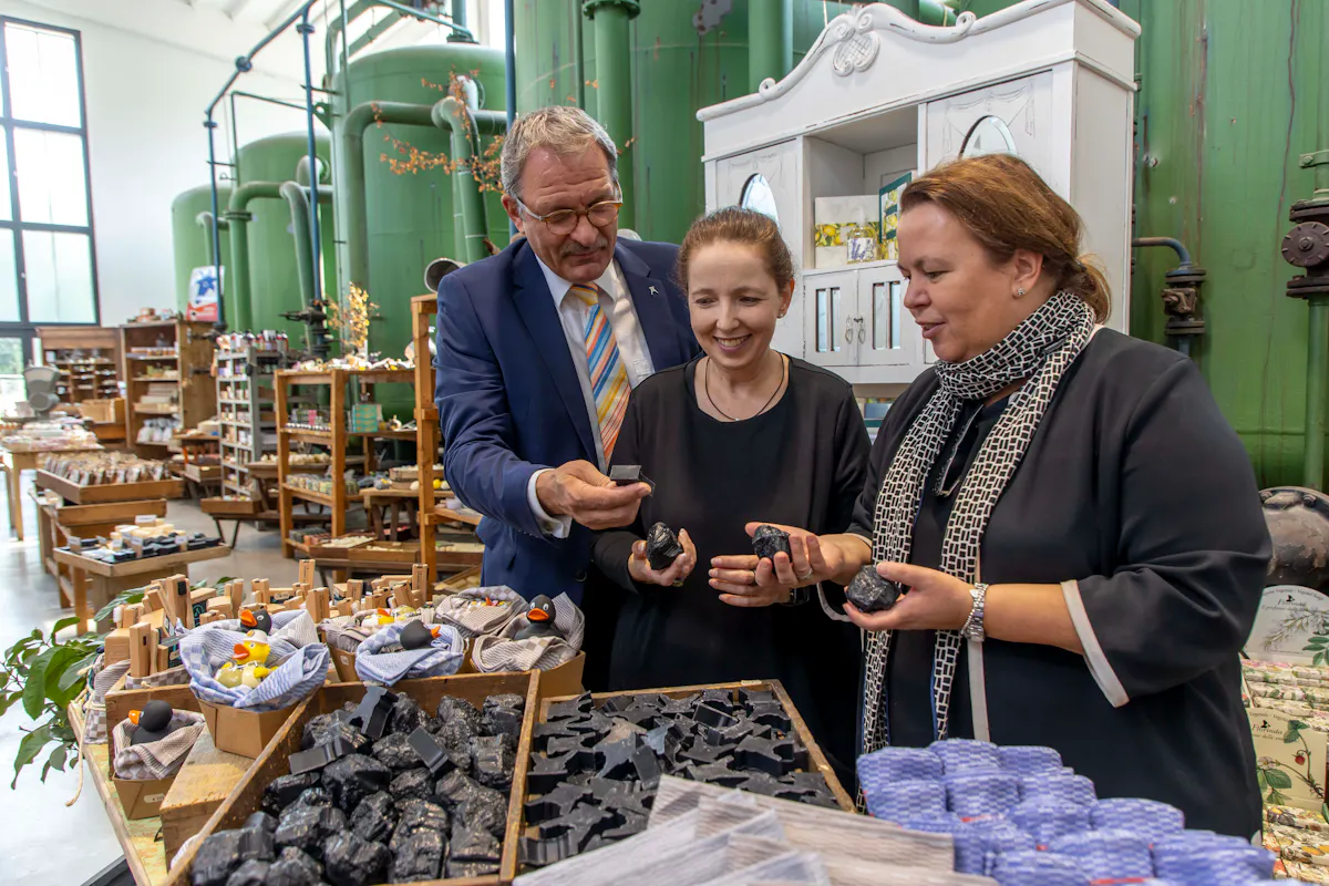 In einem liebevoll dekorierten Verkaufsraum mit industriellem Charme stehen drei Personen gemeinsam an einem Verkaufsstand. Links ein Mann im blauen Anzug mit bunter Krawatte, in der Mitte eine Frau mit zurückgebundenem Haar in schwarzem Oberteil, rechts eine Frau mit braunen Haaren, dunkler Kleidung und gemustertem Schal. Alle drei halten schwarze Seifenstücke in der Hand, die wie Kohlebrocken aussehen. Auf dem Tisch vor ihnen sind zahlreiche solcher Seifenstücke ausgestellt, daneben auch kleine Körbe mit Gummienten. Im Hintergrund stehen grüne industrielle Tanks, alte Möbelstücke und Regale mit weiteren Produkten. Die Stimmung ist freundlich und interessiert.
