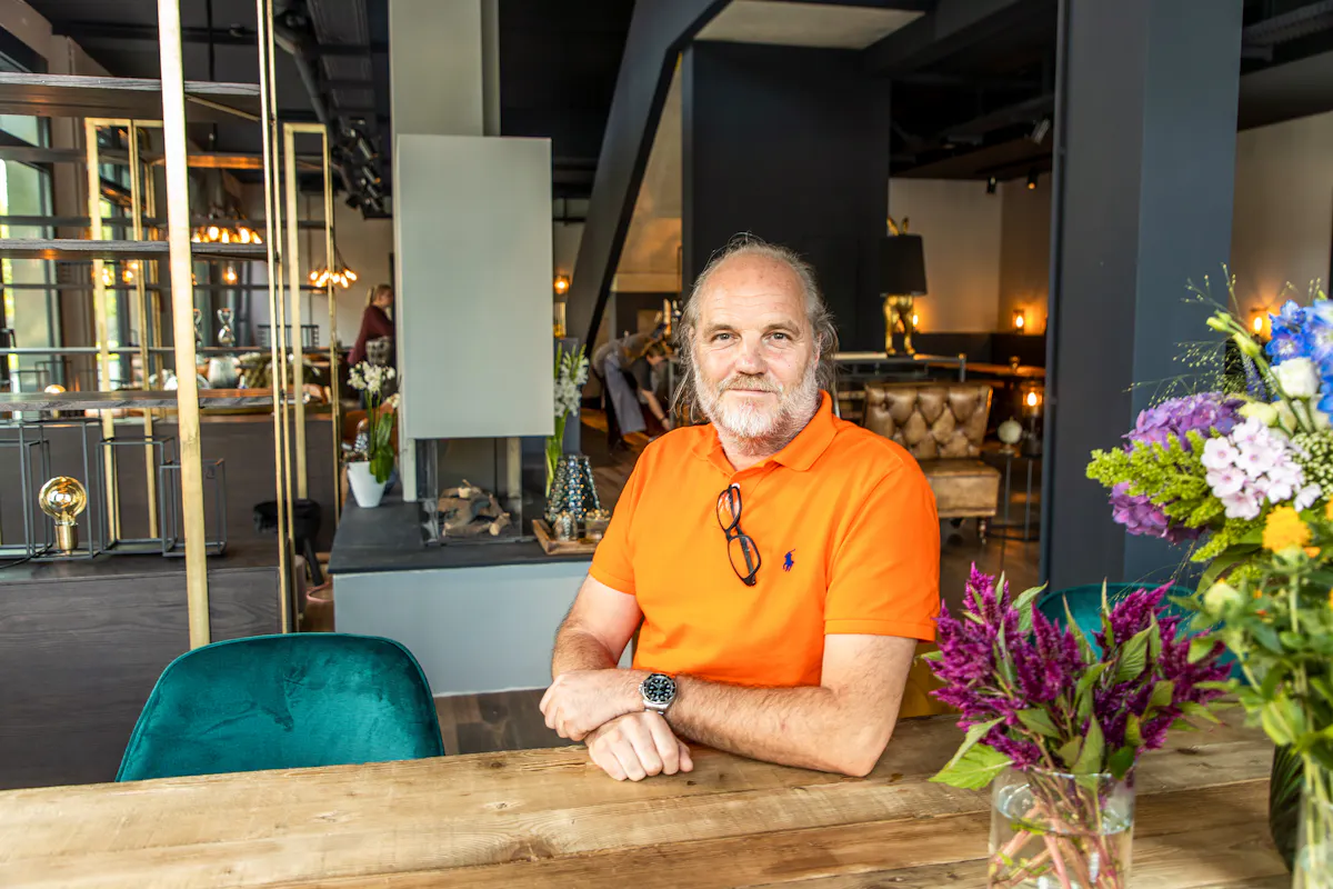 Ein älterer Mann mit grauem Haar und Bart sitzt entspannt an einem Holztisch in einem stilvoll eingerichteten, modernen Innenraum. Er trägt ein leuchtend orangefarbenes Poloshirt und hat eine Brille am Kragen sowie eine Armbanduhr am Handgelenk. Im Vordergrund stehen zwei bunte Blumensträuße in Glasvasen. Im Hintergrund sind elegante Sitzmöbel, dezente Beleuchtung und dekorative Elemente wie ein Kamin und Regale zu sehen. Die Atmosphäre wirkt gemütlich und hochwertig.