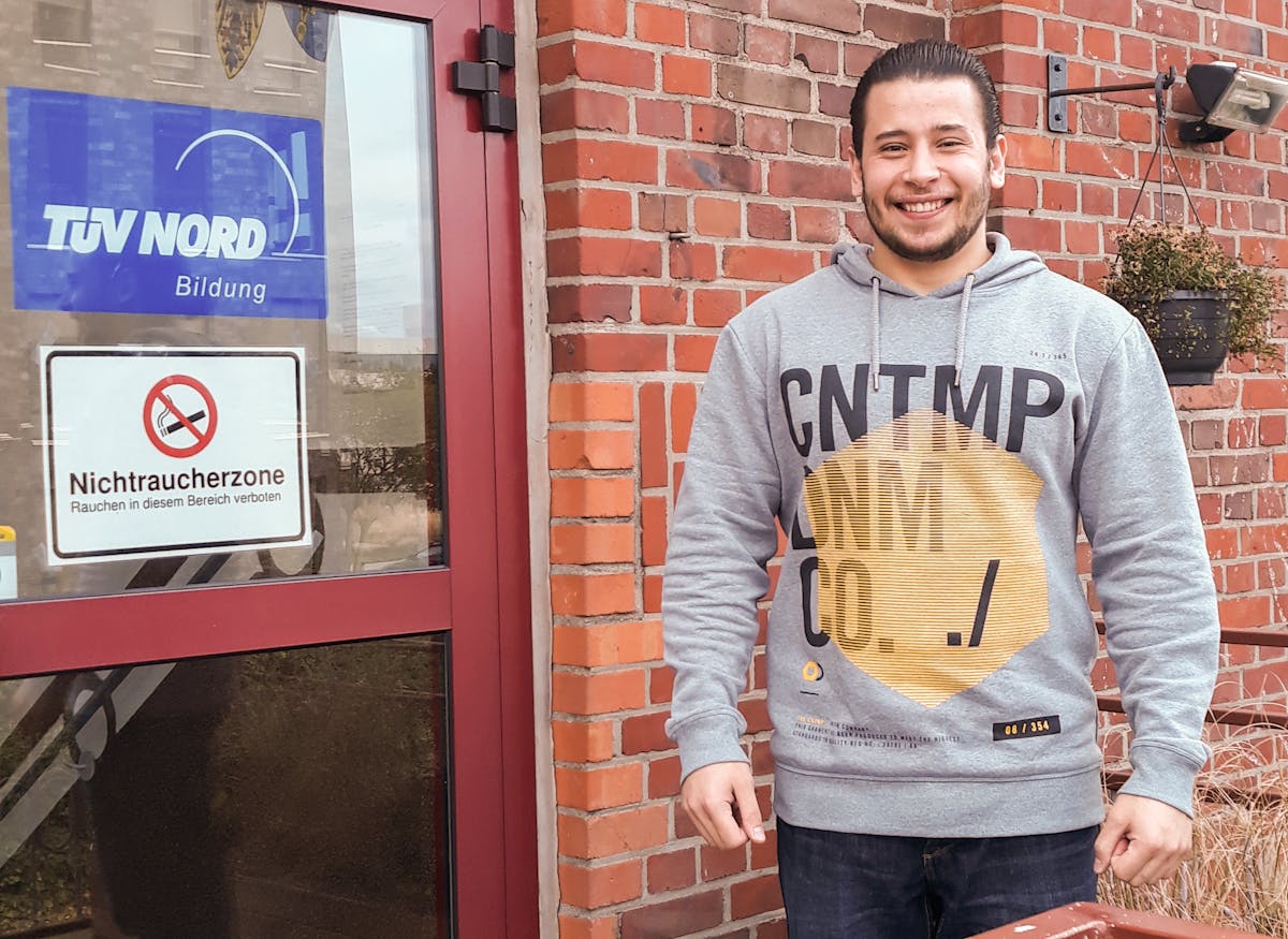 Ein junger Mann mit dunklen, zurückgekämmten Haaren und einem breiten Lächeln steht vor einem Backsteingebäude. Er trägt einen grauen Kapuzenpullover mit einem auffälligen gelben und schwarzen Aufdruck und eine dunkle Jeans. Links neben ihm befindet sich eine Glastür mit Aufklebern, darunter ein Schild mit der Aufschrift TÜV NORD Bildung sowie ein Nichtraucherschild. Rechts an der Wand hängt ein Blumenkorb mit einer Pflanze. Die Szene wirkt freundlich und einladend.