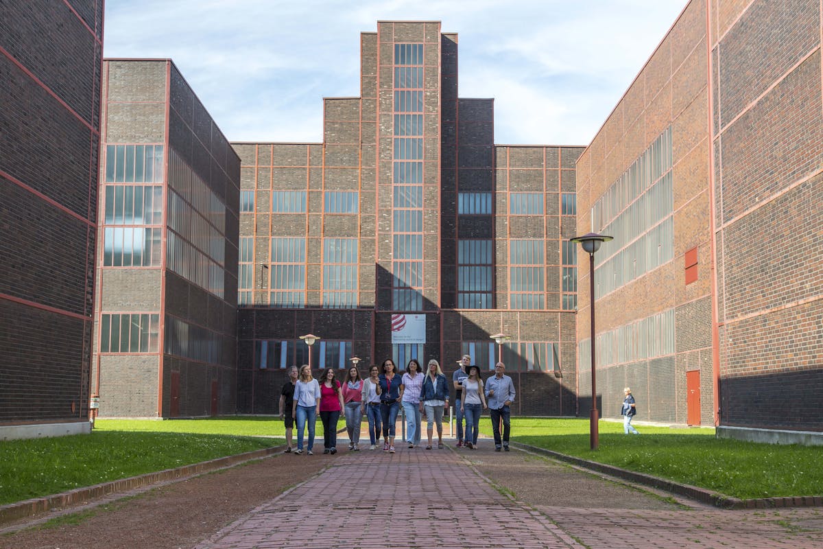 Eine Gruppe von zwölf Menschen geht in lockerer Formation auf einem gepflasterten Weg auf ein modernes Backsteingebäude mit vielen hohen Fenstern zu. Das Gebäude besitzt eine monumentale, symmetrische Architektur im Stil der Neuen Sachlichkeit mit vertikal gestaffelten Fassadenelementen. Rechts und links vom Weg befindet sich Rasenfläche. Die Szene spielt bei sonnigem Wetter auf dem Gelände des Welterbes Zollverein in Essen.