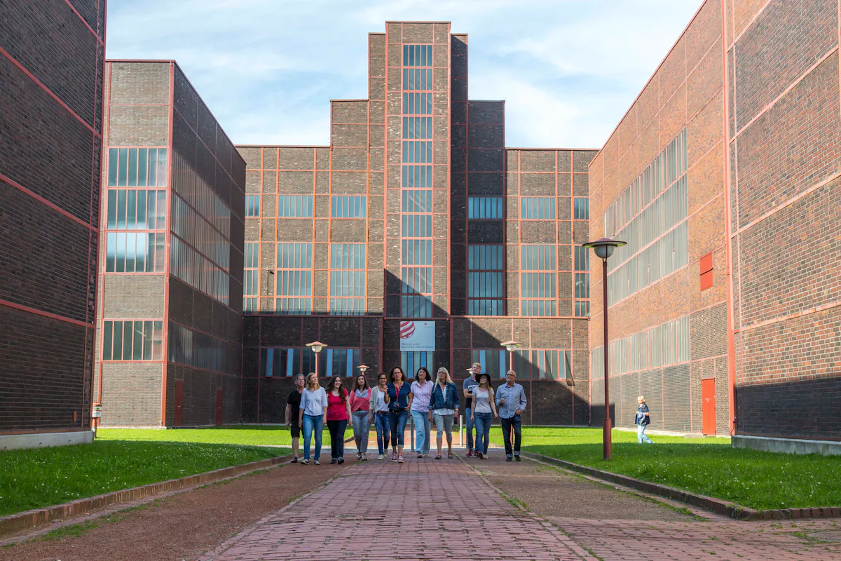 Eine Gruppe von zwölf Menschen geht in lockerer Formation auf einem gepflasterten Weg auf ein modernes Backsteingebäude mit vielen hohen Fenstern zu. Das Gebäude besitzt eine monumentale, symmetrische Architektur im Stil der Neuen Sachlichkeit mit vertikal gestaffelten Fassadenelementen. Rechts und links vom Weg befindet sich Rasenfläche. Die Szene spielt bei sonnigem Wetter auf dem Gelände des Welterbes Zollverein in Essen.