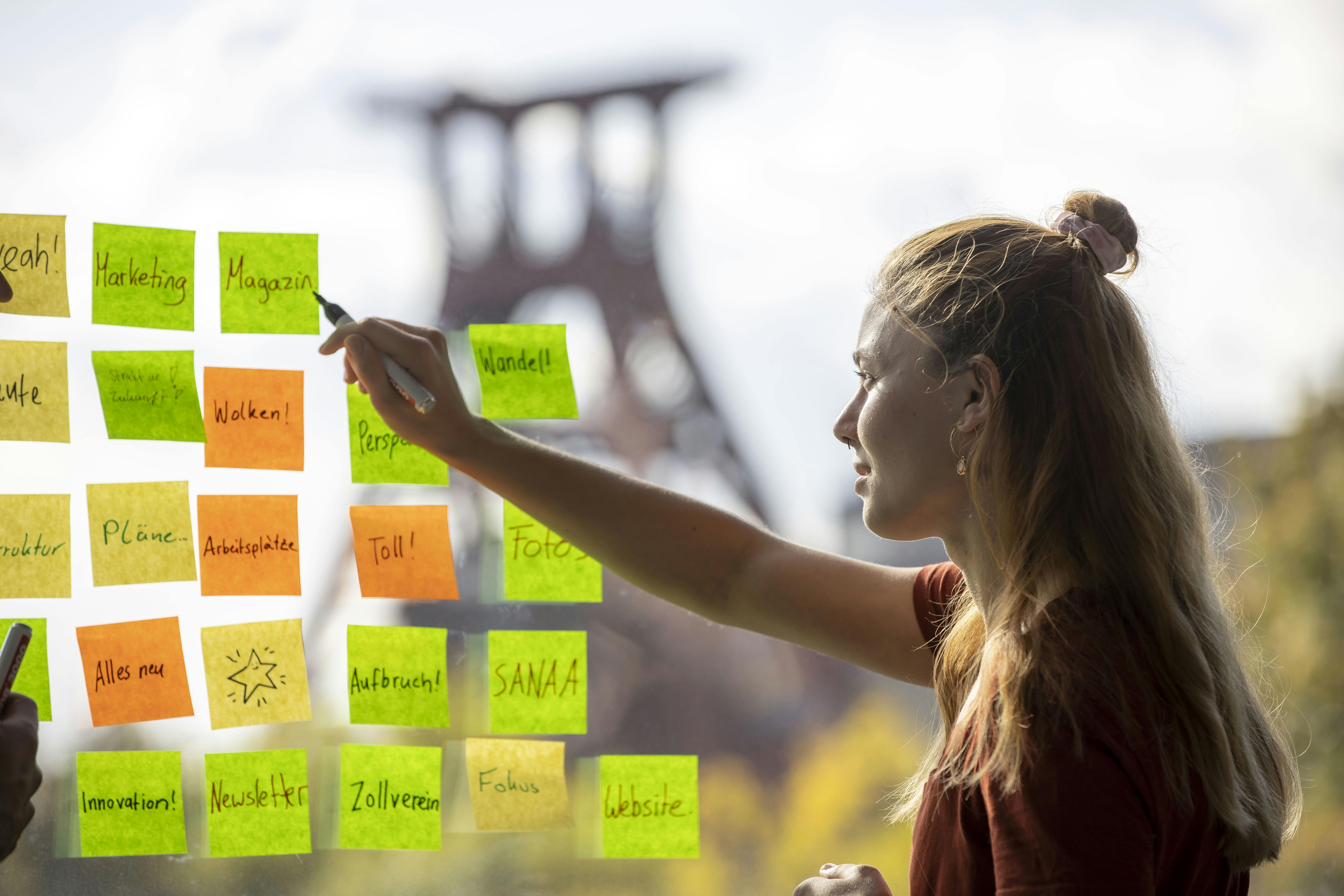 Eine junge Frau steht vor einem Fenster, an dem zahlreiche bunte Haftnotizen kleben. Sie schreibt mit einem Stift auf einen grünen Zettel, während im Hintergrund das markante Fördergerüst der Zeche Zollverein verschwommen zu erkennen ist. Die Notizen enthalten Begriffe wie Marketing, Wandel, Pläne, Website, Perspektive und Aufbruch. Die Szene wirkt kreativ und brainstormartig, vermutlich in einem Workshop- oder Projektrahmen. Die Sonne scheint, was für eine helle, freundliche Arbeitsatmosphäre sorgt.