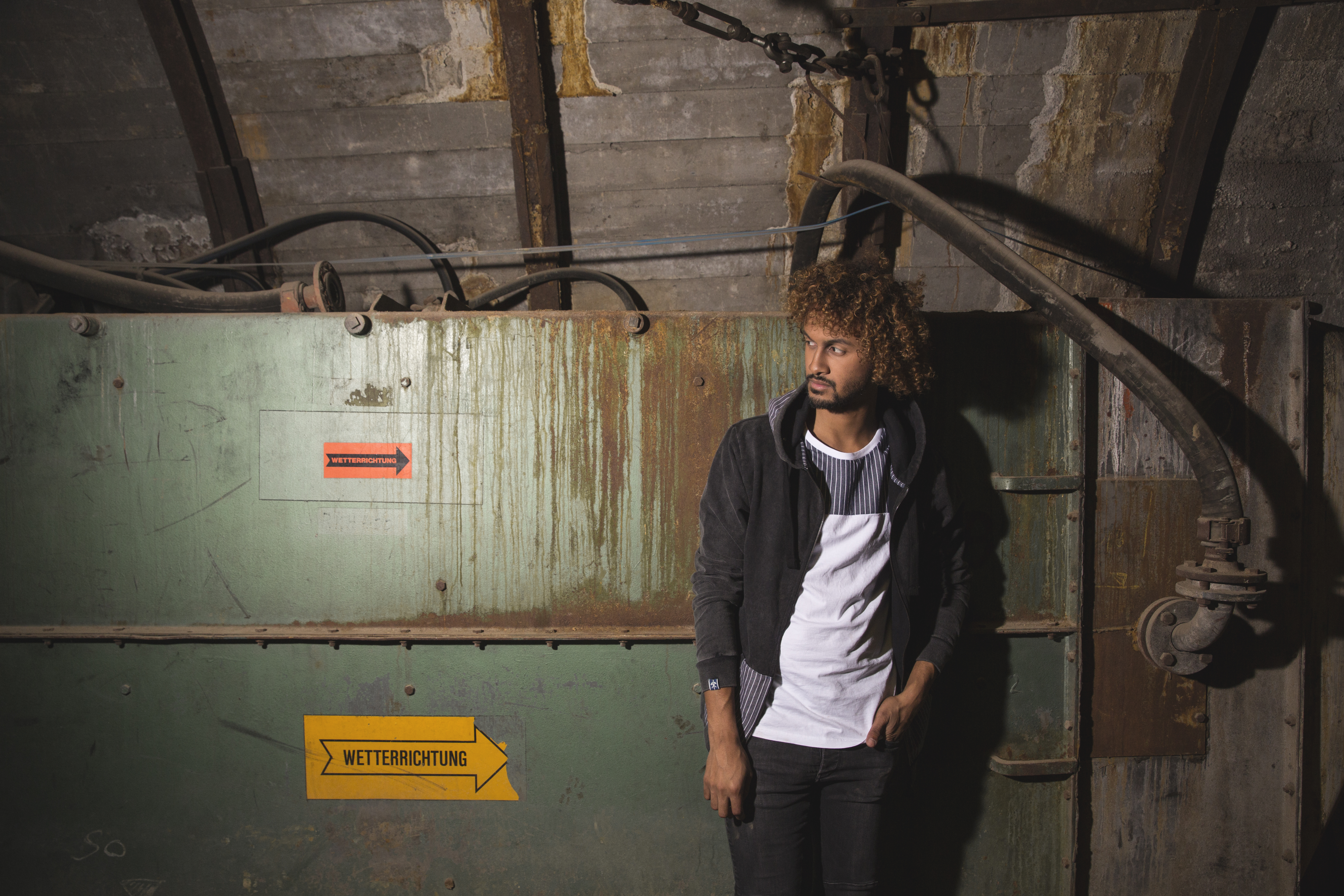 Ein junger Mann mit lockigem Haar und Bart steht in einem dunklen, industriellen Raum vor einer alten, grünlich verfärbten Metalltür oder Belüftungsklappe. Er trägt ein weißes T-Shirt, ein schwarz-graues Hemd darunter und eine schwarze Kapuzenjacke. Über ihm verlaufen Rohre und Kabel. Auf der Tür sind zwei Schilder mit der Aufschrift Wetterrichtung und einem Richtungspfeil zu sehen – eines gelb, eines orange. Die Szenerie wirkt wie in einem ehemaligen Bergwerksstollen mit starkem Licht-Schatten-Kontrast.