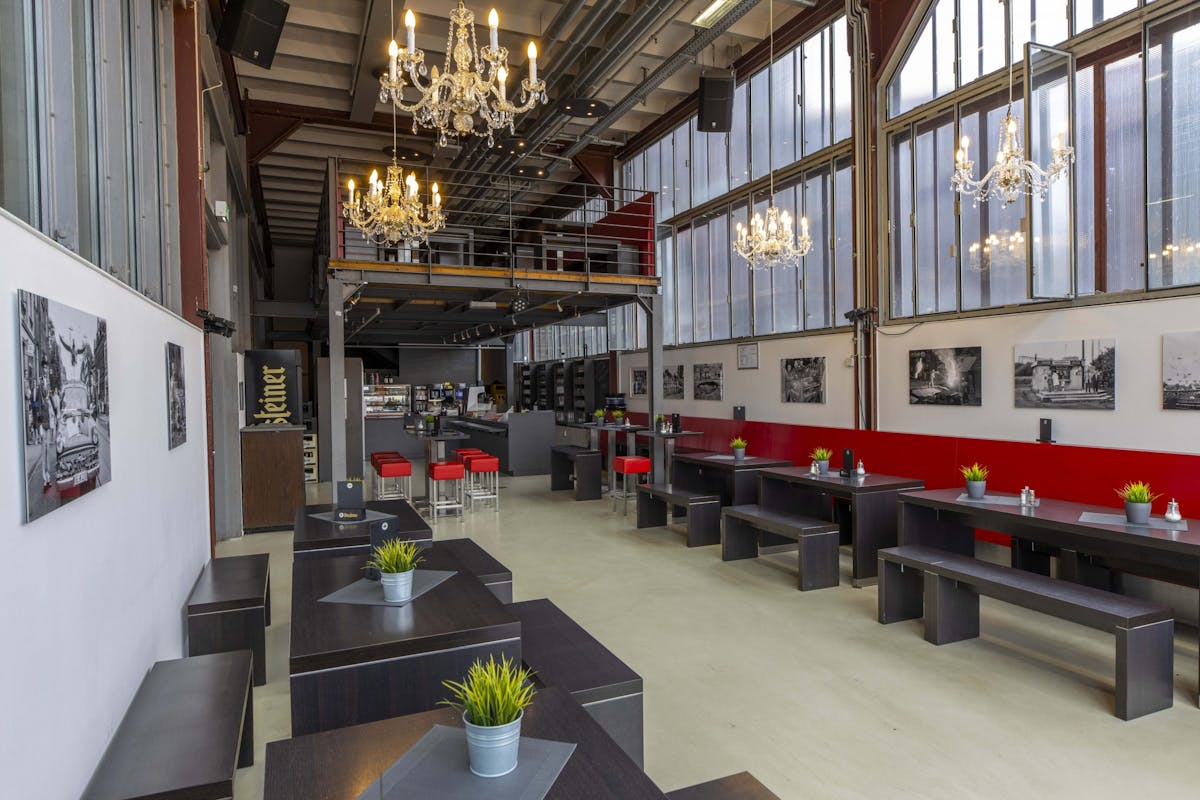 Das Bild zeigt den Innenraum eines modern eingerichteten Gastraums in einer Industriehalle mit hohen Decken und großen Fensterflächen. Entlang der rechten Wand stehen dunkle Holzbänke und -tische mit kleinen Dekopflanzen in Metalltöpfen. An der Wand darüber hängen Schwarz-Weiß-Fotografien mit industriellen oder historischen Motiven. In der Mitte des Raumes befinden sich weitere Sitzgelegenheiten sowie eine Bar mit roten Barhockern. Von der Decke hängen mehrere opulente Kronleuchter, die einen stilvollen Kontrast zur industriellen Umgebung bilden. Im Hintergrund führt eine Treppe zu einer Empore mit zusätzlichem Raum. Die Atmosphäre wirkt freundlich, aufgeräumt und einladend.
