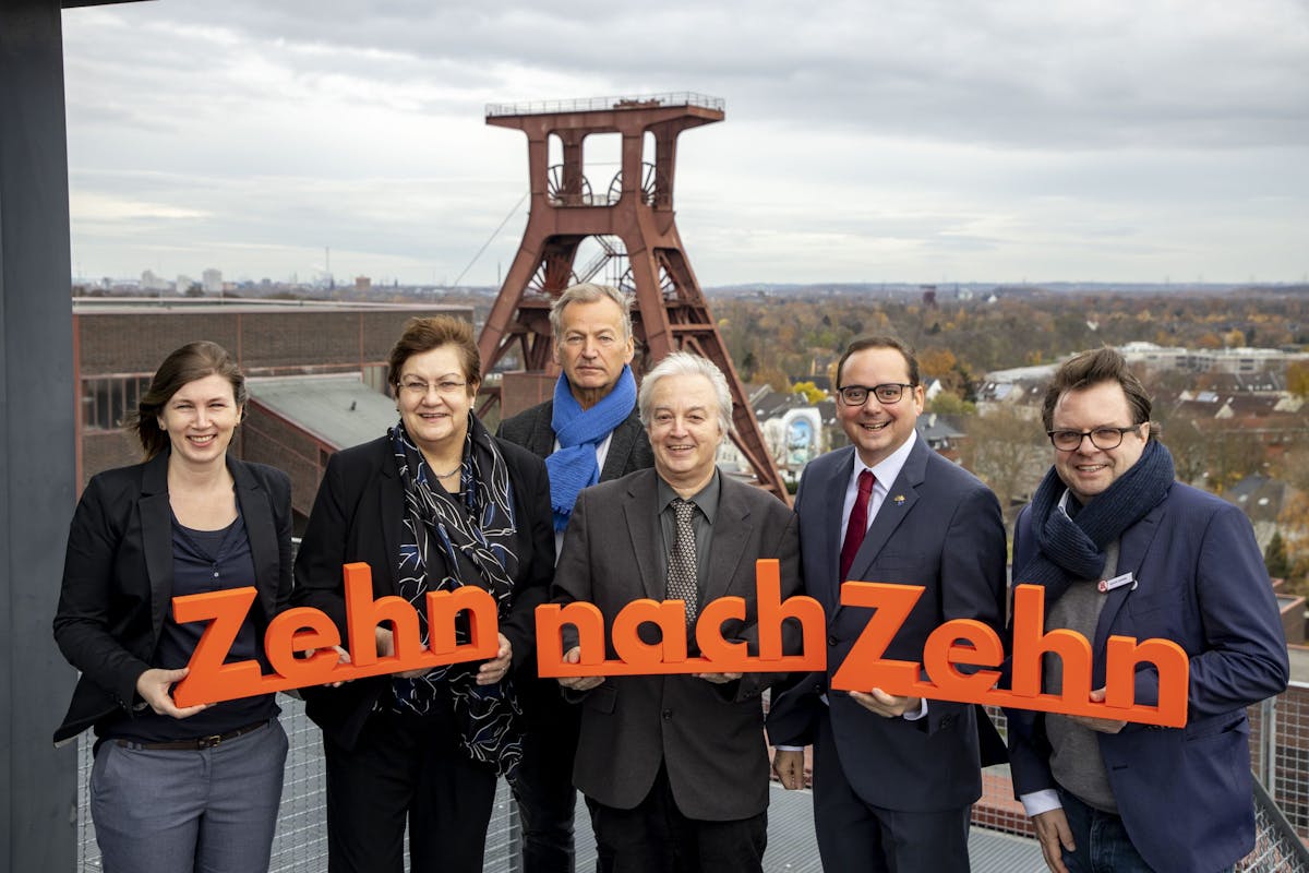 Das Bild zeigt sechs Personen – drei Frauen und drei Männer – die nebeneinander auf einer Aussichtsplattform vor dem markanten Fördergerüst der Zeche Zollverein in Essen stehen. Alle lächeln in die Kamera und tragen formelle Kleidung; zwei Männer tragen zusätzlich blaue Schals. Die Gruppe hält gemeinsam ein großes, leuchtend orangefarbenes Schriftobjekt aus Einzelsegmenten, das den Schriftzug Zehn nach Zehn bildet. Die Buchstaben sind dreidimensional und in einer serifenlosen, modernen Schriftart gestaltet. Jedes Wort ist einzeln gehalten – das erste Zehn links, das Wort nach in der Mitte, das zweite Zehn rechts. Die Buchstaben sind gleichmäßig groß, sauber geschnitten und werden von den Personen jeweils mit beiden Händen gehalten oder abgestützt. Im Hintergrund ist ein herbstlich gefärbter Waldgürtel sowie ein weiter Blick über die Stadt Essen unter einem wolkigen Himmel zu sehen. Die Szene wirkt freundlich, offiziell und ist vermutlich Teil einer Jubiläums- oder PR-Aktion.