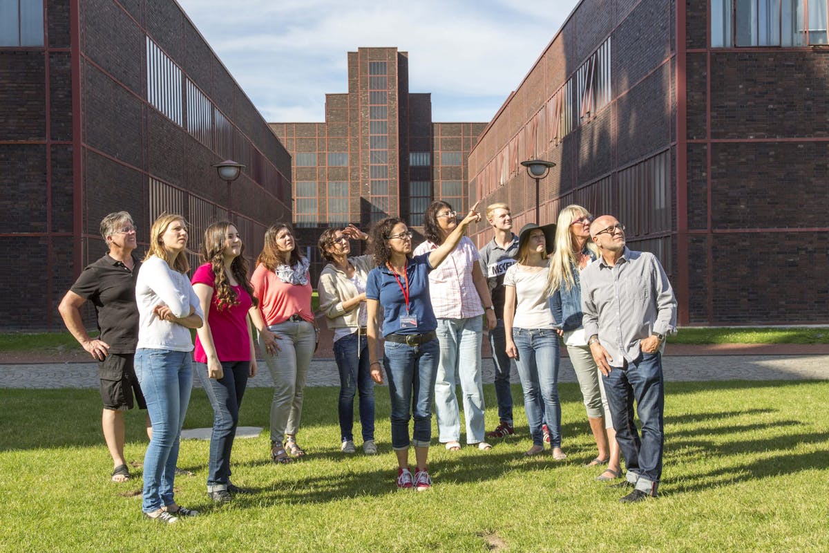 Eine Gruppe von etwa 14 Erwachsenen steht bei sonnigem Wetter auf einer Rasenfläche zwischen zwei symmetrisch verlaufenden Backsteingebäuden auf dem Gelände des UNESCO-Welterbes Zollverein. Die Personen blicken interessiert nach oben, eine Frau in der Mitte zeigt mit ausgestrecktem Arm auf etwas am Himmel oder an einem der Gebäude. Die Szene wirkt wie eine geführte Tour oder ein gemeinsamer Rundgang durch das Industriegelände. Im Hintergrund ist ein zentrales Gebäude mit rot gerahmten Fenstern sichtbar, das perspektivisch den Blickpunkt der Architektur bildet.