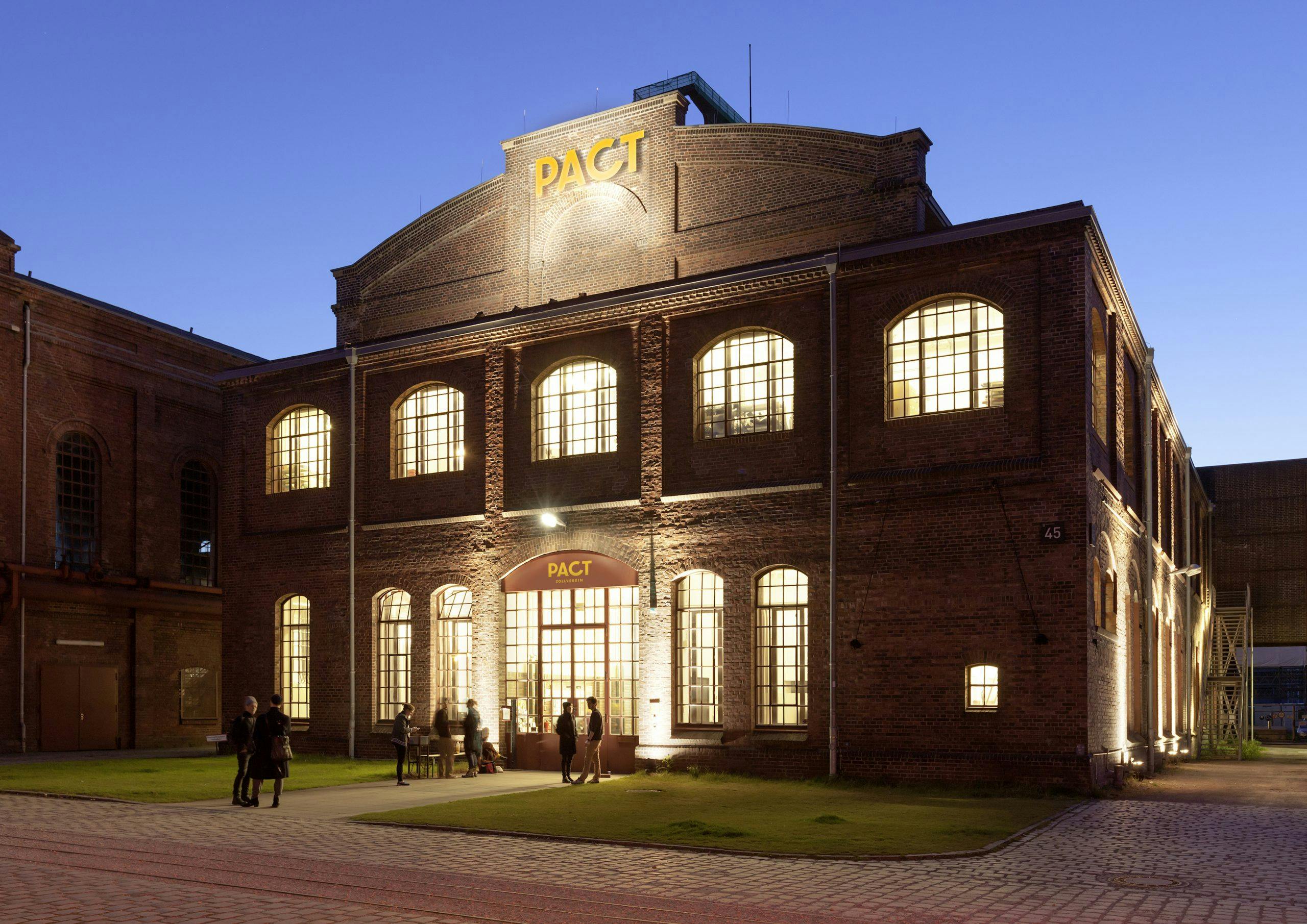 Backstein-Gebäude des Kulturzentrums PACT Zollverein in der Dämmerung, hell beleuchtet von innen durch große, hohe Fenster. Über dem Eingang ist der gelbe Schriftzug PACT deutlich sichtbar. Vor dem Eingang versammeln sich mehrere Personen, manche stehen im Gespräch, andere betreten das Gebäude. Das Gebäude wirkt historisch-industriell mit moderner Nutzung, und die Szene strahlt eine einladende, kulturelle Atmosphäre aus.