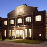 Backstein-Gebäude des Kulturzentrums PACT Zollverein in der Dämmerung, hell beleuchtet von innen durch große, hohe Fenster. Über dem Eingang ist der gelbe Schriftzug PACT deutlich sichtbar. Vor dem Eingang versammeln sich mehrere Personen, manche stehen im Gespräch, andere betreten das Gebäude. Das Gebäude wirkt historisch-industriell mit moderner Nutzung, und die Szene strahlt eine einladende, kulturelle Atmosphäre aus.