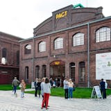 Das Bild zeigt das rote Backsteingebäude von PACT Zollverein, einem Zentrum für Tanz, Performance, Kunst und Theater auf dem UNESCO-Welterbe Zollverein. Auf dem Gebäude prangt oben deutlich sichtbar in gelben Großbuchstaben das Wort PACT. Darunter befindet sich ein Eingang mit einem Vordach, über dem ebenfalls PACT in gelber Schrift steht. An der Fassade rechts ist ein großes weißes Plakat mit dem Programm oder Veranstaltungshinweisen angebracht. Vor dem Gebäude stehen und unterhalten sich mehrere Personen, einige sehen sich die Plakate an. Der Vorplatz ist mit Kopfsteinpflaster ausgelegt und teilweise begrünt. Ein kleiner Hund ist im Vordergrund zu sehen. Die Atmosphäre wirkt ruhig und einladend, als ob die Gäste auf eine Veranstaltung warten oder gerade ankommen.