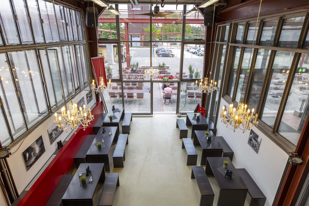 Innenaufnahme eines hohen, lichtdurchfluteten Gastraums mit bodentiefen Fenstern auf beiden Seiten und einem zentralen Ausgang zur Außenterrasse mit Holzbänken. Im Raum stehen dunkle Holztische mit passenden Bänken, auf denen jeweils kleine grüne Pflanzen in Töpfen platziert sind. Zwei große Kronleuchter hängen von der Decke und verleihen dem Raum einen eleganten Akzent. An den weißen Wänden sind Schwarz-Weiß-Fotografien angebracht, eine Seite des Raumes wird zusätzlich durch eine rote Wandverkleidung strukturiert.
