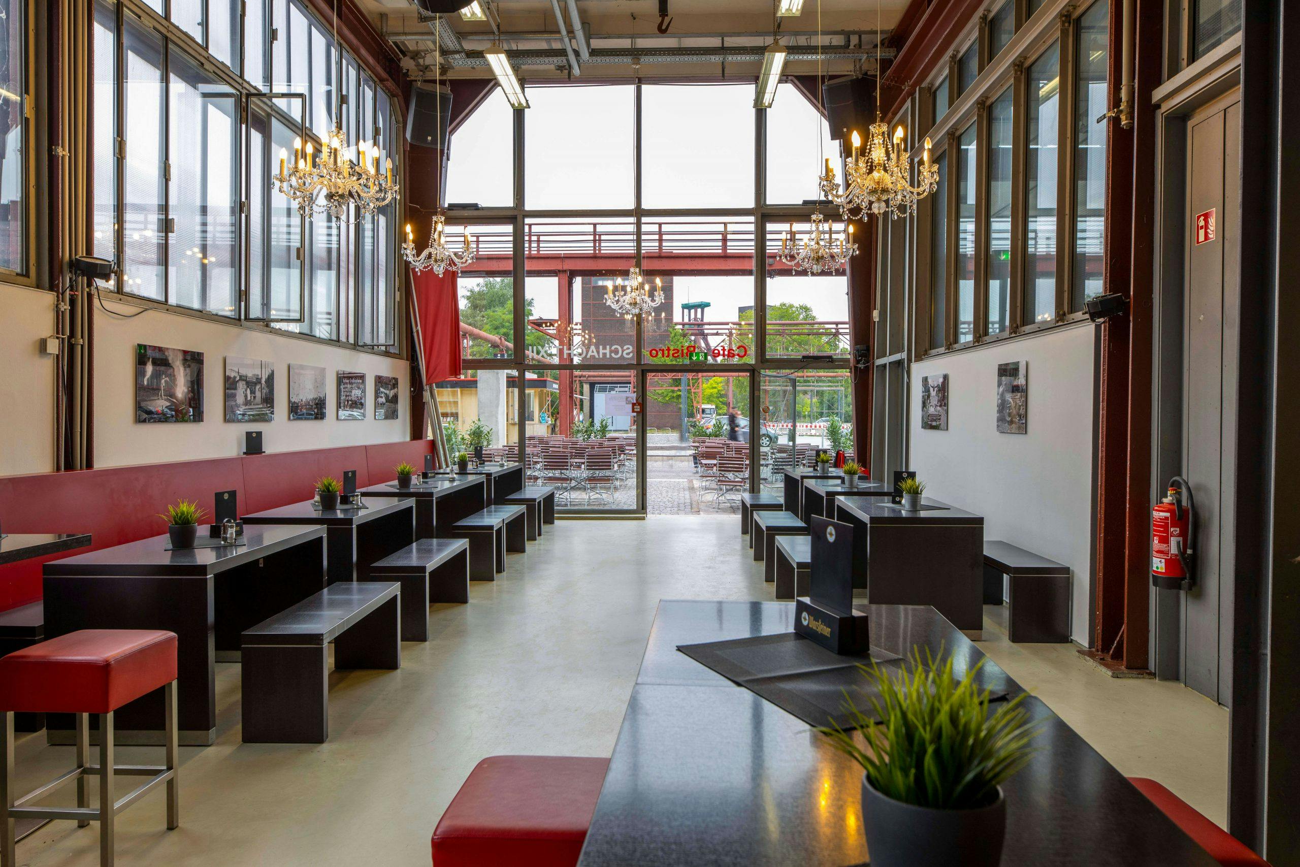 Innenaufnahme eines modern gestalteten Cafés mit industriellem Charme und hoher Decke. Der Raum ist länglich, mit bodentiefen Fenstern an beiden Seiten, durch die viel Tageslicht fällt. An den Seitenwänden stehen schwarze Tische mit passenden Bänken, dekoriert mit kleinen grünen Kunstpflanzen in silbernen Töpfen. Links zieht sich eine rote Rückenlehne entlang der Wand, darüber hängen gerahmte Schwarz-Weiß-Fotografien. Auffällig sind mehrere große Kronleuchter, die elegant von der Decke hängen und dem Raum einen stilvollen Kontrast zur industriellen Architektur verleihen. Im Hintergrund führt eine große Glasfront mit Tür nach draußen zu einem gepflasterten Bereich mit weiteren Sitzgelegenheiten. Auf der Tür steht in roter Schrift Café Bistro Schacht 12 geschrieben.