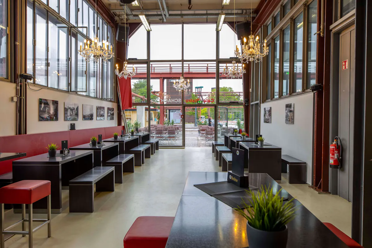 Innenaufnahme eines modern gestalteten Cafés mit industriellem Charme und hoher Decke. Der Raum ist länglich, mit bodentiefen Fenstern an beiden Seiten, durch die viel Tageslicht fällt. An den Seitenwänden stehen schwarze Tische mit passenden Bänken, dekoriert mit kleinen grünen Kunstpflanzen in silbernen Töpfen. Links zieht sich eine rote Rückenlehne entlang der Wand, darüber hängen gerahmte Schwarz-Weiß-Fotografien. Auffällig sind mehrere große Kronleuchter, die elegant von der Decke hängen und dem Raum einen stilvollen Kontrast zur industriellen Architektur verleihen. Im Hintergrund führt eine große Glasfront mit Tür nach draußen zu einem gepflasterten Bereich mit weiteren Sitzgelegenheiten. Auf der Tür steht in roter Schrift Café Bistro Schacht 12 geschrieben.