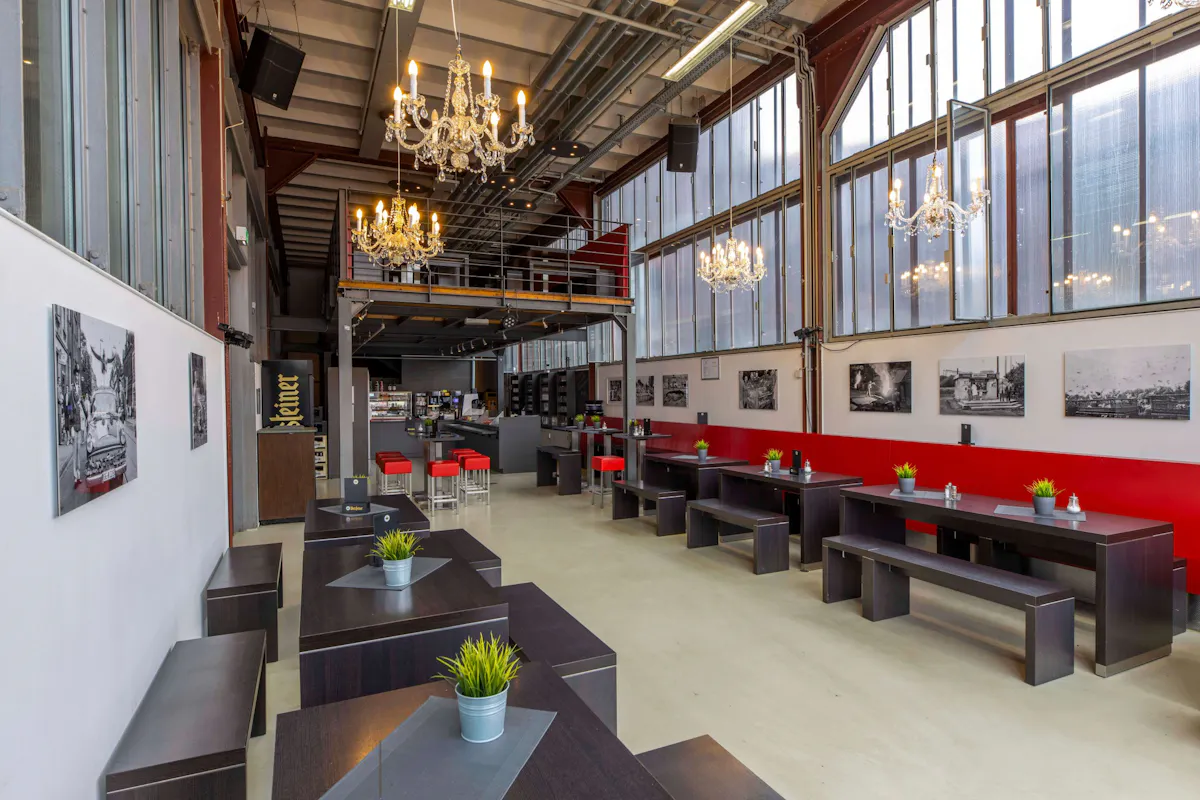 Innenansicht eines modern eingerichteten Cafés mit industriellem Design und hoher Decke. Der Raum ist langgestreckt mit großen Fensterflächen an beiden Seiten, die viel Tageslicht hereinlassen. Es gibt mehrere dunkle Holztische mit passenden Bänken, auf denen kleine grüne Kunstpflanzen in silbernen Töpfen und Menükarten platziert sind. Die rechte Wand ist durchgehend mit einer roten Rückenlehne versehen und mit Schwarz-Weiß-Fotografien dekoriert. An der Decke hängen elegante Kronleuchter, die einen stilvollen Kontrast zur industriellen Einrichtung mit sichtbaren Leitungen und Stahlträgern bilden. Im Hintergrund befindet sich eine Theke mit Kaffeemaschine und Regalen, darüber eine offene Empore mit zusätzlichem Sitzbereich. Links neben der Theke ist ein Getränkekühlschrank zu sehen.