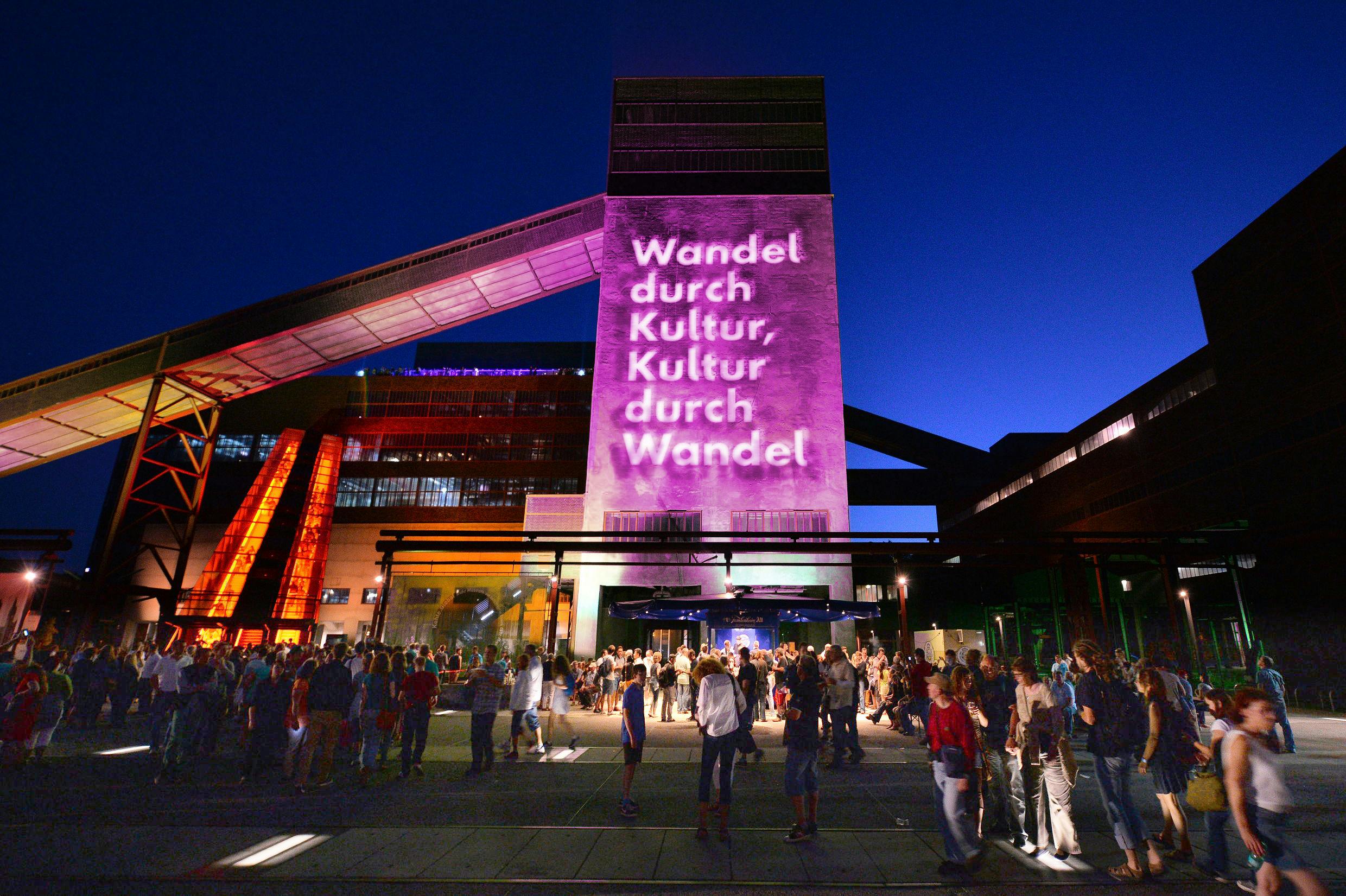 Abendaufnahme des Zollverein-Geländes mit dem markanten Kokskohlenbunker und der Rolltreppe, die orange beleuchtet ist. Auf einem zentralen Turm ist mit pink-violettem Licht der Schriftzug Wandel durch Kultur, Kultur durch Wandel projiziert. Vor dem Gebäude versammeln sich zahlreiche Menschen bei einer Veranstaltung, der Himmel ist bereits dunkelblau. Das Bild vermittelt eine lebendige Atmosphäre eines Kulturereignisses bei Nacht.