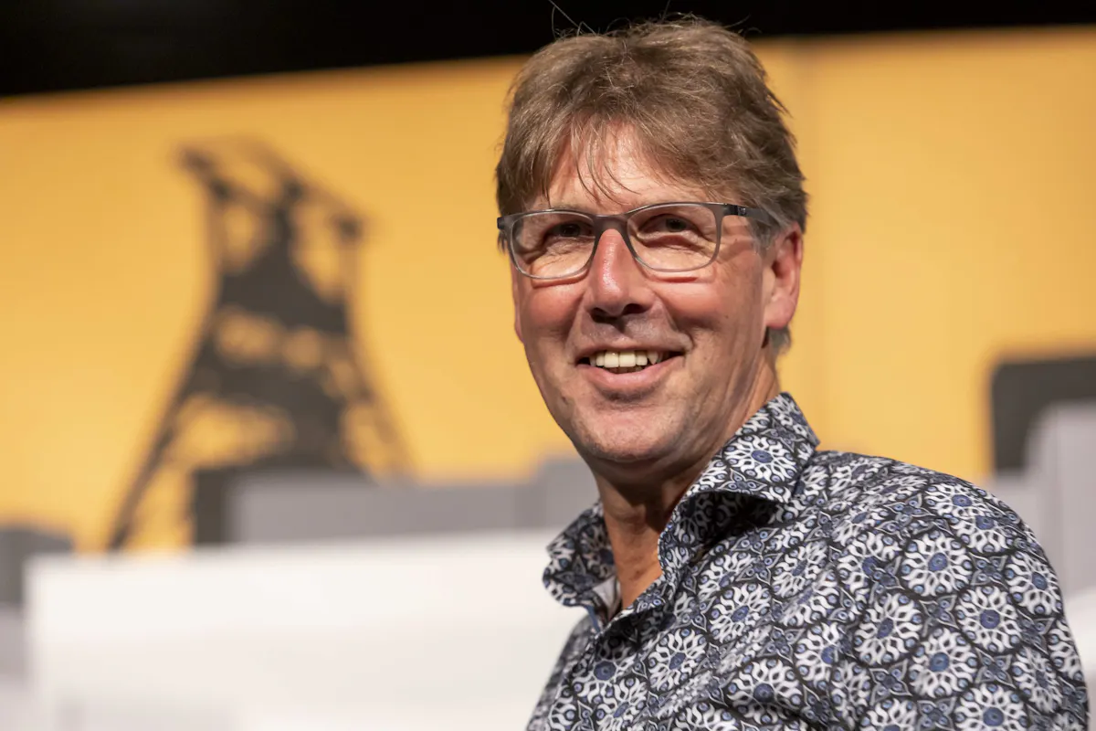 Ein lächelnder Mann mittleren Alters, der Moderator Peter Großmann, mit bräunlichen Haaren und Brille steht vor einem gelb-schwarzen Hintergrund, auf dem schemenhaft das markante Fördergerüst des Welterbes Zollverein zu erkennen ist. Der Mann trägt ein gemustertes Hemd in Blau- und Weißtönen und blickt direkt in die Kamera. Die Szene wirkt freundlich und professionell.