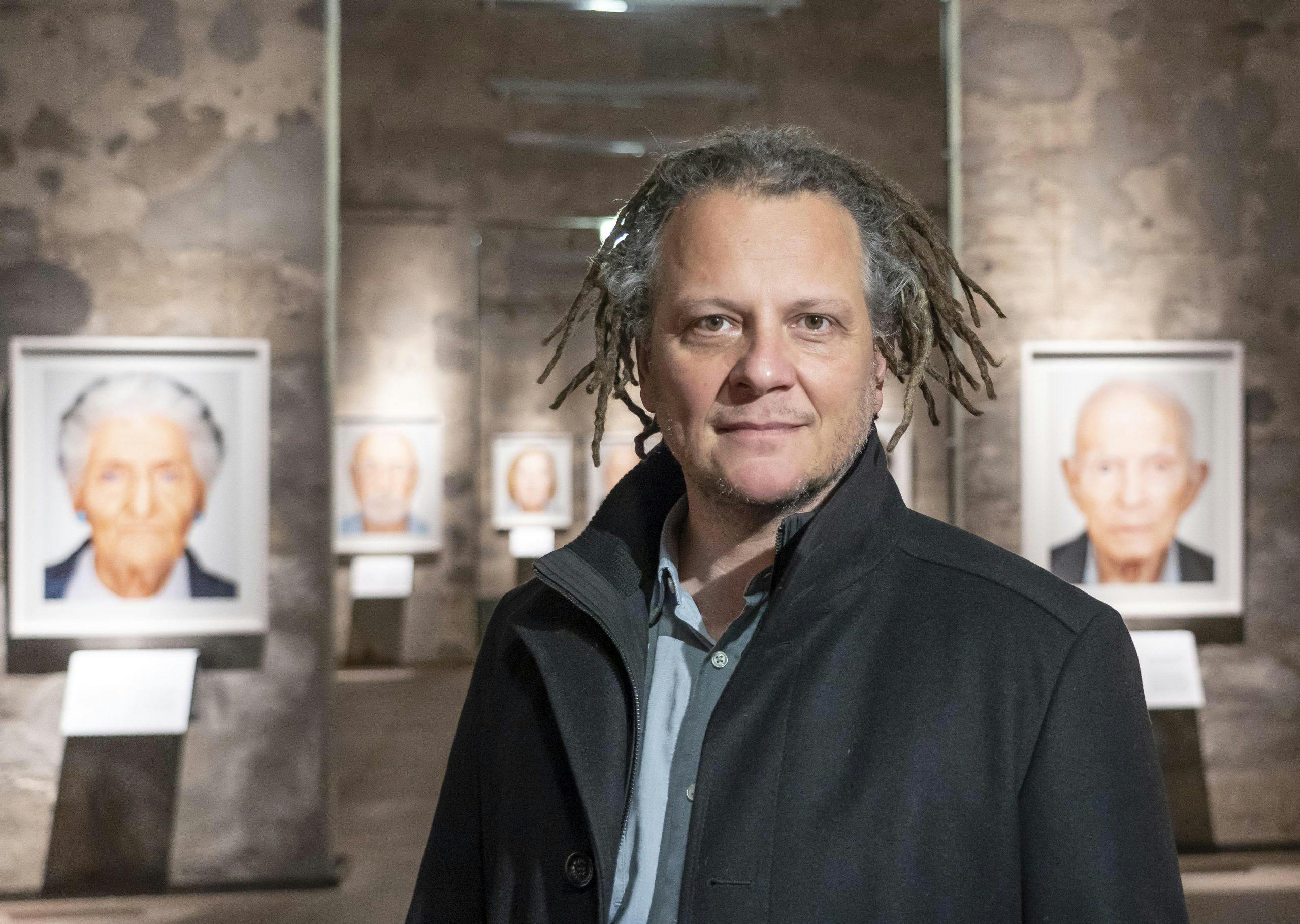 Ein Mann, der Fotograf Martin Schoeller, mit mittellangen, lockigen Dreadlocks, hellblauem Hemd und schwarzem Mantel steht in einer Galerie vor einer Wand aus rauem Stein. Im Hintergrund sind großformatige Porträtfotos älterer Menschen in weißen Rahmen ausgestellt, die gleichmäßig an der Wand hängen und von kleinen Infotafeln begleitet werden. Die Lichtstimmung ist warm und die Atmosphäre ruhig und konzentriert.