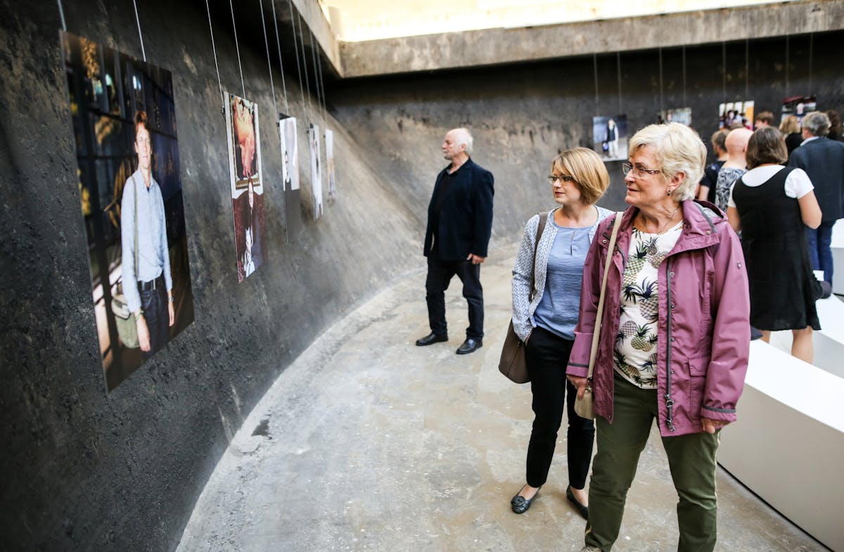 Mehrere Besucherinnen und Besucher betrachten eine Fotoausstellung in einem abgerundeten, rauen Raum mit Betonwänden. Die großformatigen Porträtfotografien sind in einer Reihe aufgehängt und zeigen verschiedene Menschen. Im Vordergrund stehen zwei Frauen mittleren Alters, eine in lilafarbener Jacke, die andere in hellblauer Bluse, beide schauen interessiert auf die Bilder. Weitere Personen befinden sich im Hintergrund und bewegen sich durch die Ausstellung. Die Atmosphäre wirkt ruhig und konzentriert.