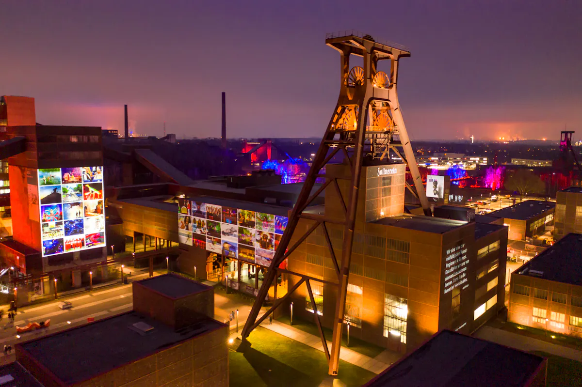 Nachtaufnahme des Welterbes Zollverein mit stimmungsvoller Beleuchtung. Das markante Doppelbock-Fördergerüst ist angestrahlt und überragt das Ruhr Museum, dessen Fassaden mit großflächigen, leuchtenden Projektionen von Fotos und Texten bespielt werden. Auf einem Gebäude ist der Schriftzug Zollverein zu sehen. Im Hintergrund sind weitere Teile des Geländes farbig illuminiert, darunter Bäume in Lila und Blau. Der Himmel ist dunkelviolett mit einem fernen Lichtschein am Horizont. Die Szene wirkt festlich und lebendig.