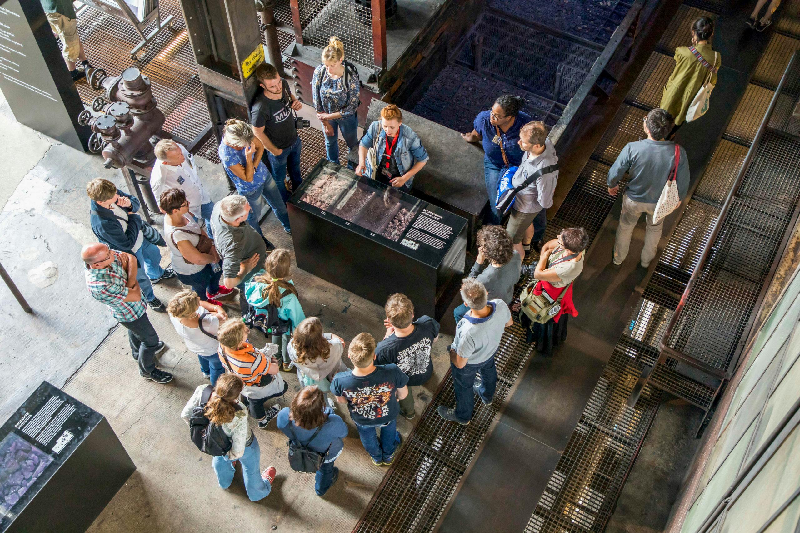 Eine Gruppe von Menschen steht in einem Industriebau auf dem Gelände der Zeche Zollverein und hört einer Gästeführerin zu. Die Gruppe ist um ein schwarzes Informationsschaukasten versammelt, das mit Kohle, Steinen und dazugehörigen Texten ausgestattet ist. Die Umgebung besteht aus industriellen Materialien wie Metallgittern, Stahlrohren und Beton, die an die frühere Nutzung als Kohlenwäsche erinnern.