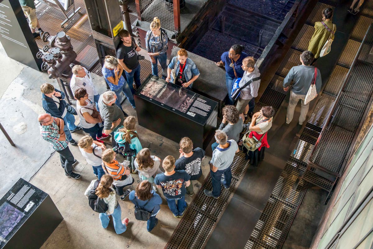 Eine Gruppe von Menschen steht in einem Industriebau auf dem Gelände der Zeche Zollverein und hört einer Gästeführerin zu. Die Gruppe ist um ein schwarzes Informationsschaukasten versammelt, das mit Kohle, Steinen und dazugehörigen Texten ausgestattet ist. Die Umgebung besteht aus industriellen Materialien wie Metallgittern, Stahlrohren und Beton, die an die frühere Nutzung als Kohlenwäsche erinnern.