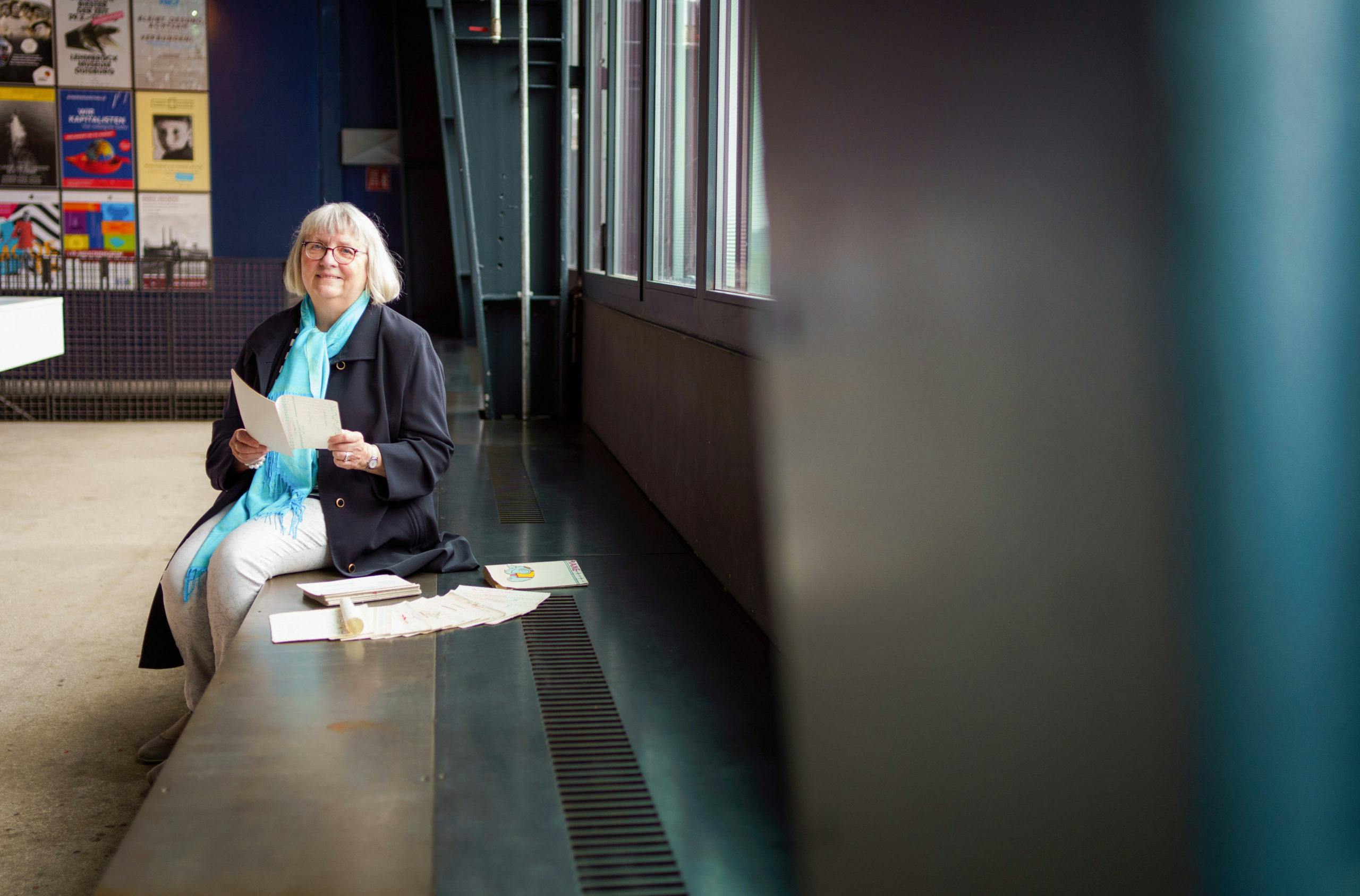 Eine ältere Frau mit weiß-grauem Haar und Brille sitzt auf einer Bank in einem modernen Gebäude mit großen Fenstern. Sie trägt einen schwarzen Mantel und einen hellblauen Schal und hält mehrere Papierseiten in der Hand. Auf der Bank vor ihr liegen weitere Blätter und ein Stempel, im Hintergrund sind bunte Plakate an der Wand zu sehen.