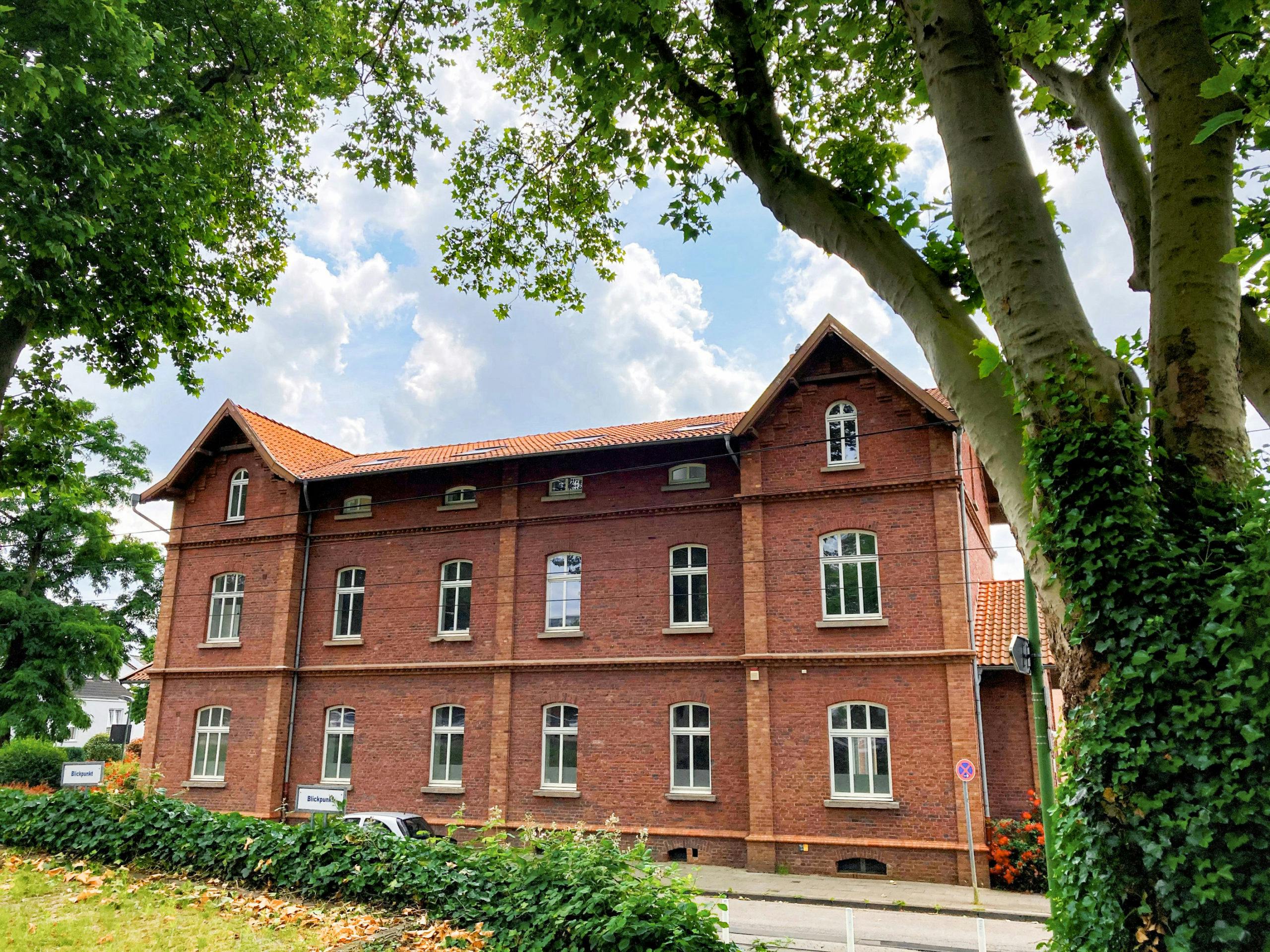 Ein zweigeschossiges Backsteingebäude mit rotem Ziegeldach steht an einer ruhigen Straße, umgeben von großen Bäumen mit dichtem Laub. Das historische Haus hat symmetrisch angeordnete weiße Fenster mit Rundbögen und kleine Giebel an den beiden Enden. Im Vordergrund wachsen Efeu und Sträucher, während rechts ein mächtiger Baum mit hellem Stamm zu sehen ist. Ein Schild mit der Aufschrift Biopark steht links am Grundstücksrand. Der Himmel ist leicht bewölkt mit sonnigen Abschnitten, was der Szene eine freundliche Atmosphäre verleiht.