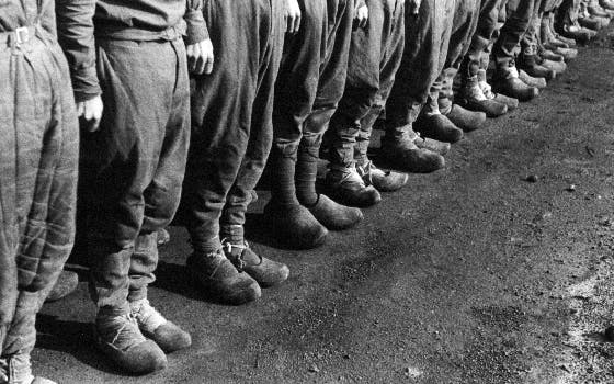 Schwarz-weiß-Foto einer Reihe von Bergleuten, die nebeneinanderstehen. Zu sehen sind ihre Beine in schmutziger Arbeitskleidung und großen Holzschuhen. Der Boden ist unbefestigt und wirkt staubig oder mit Kohlerückständen bedeckt. Insgesamt zeugt die Szene von Schmutz und Entbehrung und Zwang.