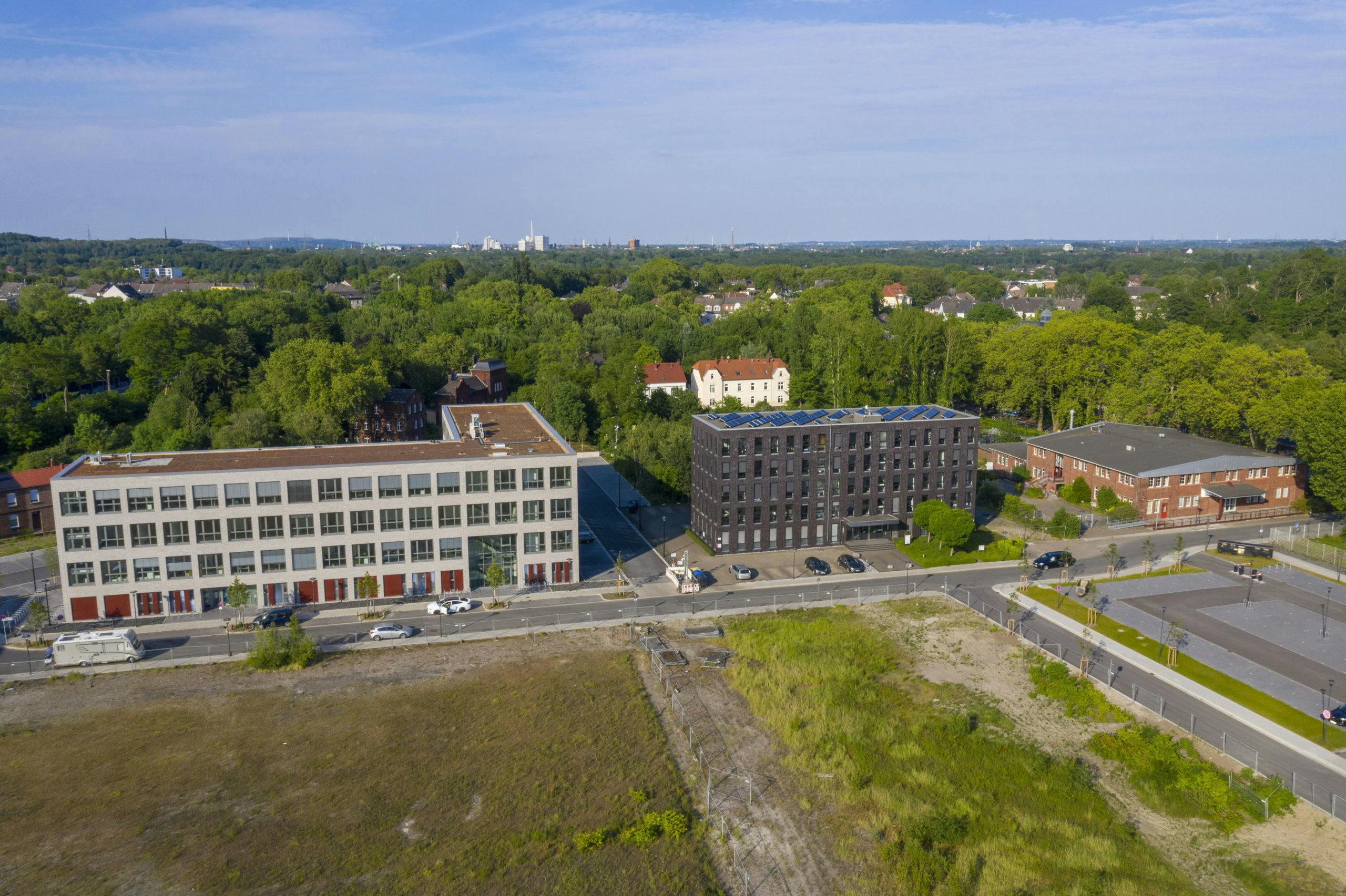 Luftaufnahme eines modernen Büroareals am Rand des Welterbes Zollverein. Im Vordergrund verläuft eine Straße mit parkenden Autos, dahinter stehen links ein helles Gebäude mit großen Fenstern und roten Türen und daneben ein dunkles Gebäude mit Solarpanelen auf dem Dach. Rechts schließt sich ein weiteres flaches Backsteingebäude an. Im Hintergrund ist ein grüner Baumbestand zu sehen, darüber erstreckt sich der Blick in die weite Stadtlandschaft.