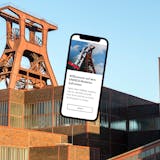 Das Bild zeigt das bekannte Doppelbock-Fördergerüst der Zeche Zollverein in Essen bei Tageslicht mit klarem Himmel im Hintergrund. Im Vordergrund ist ein überdimensionales Smartphone eingeblendet, auf dessen Bildschirm die mobile Website des UNESCO-Welterbes Zollverein geöffnet ist. Auf dem Display ist ein Willkommensgruß zu lesen, der Besucher zur Entdeckung der Welt von Kohle, Koks, Kunst und Kultur im Ruhrgebiet einlädt. Die Gestaltung verbindet historische Industriekultur mit moderner digitaler Information.