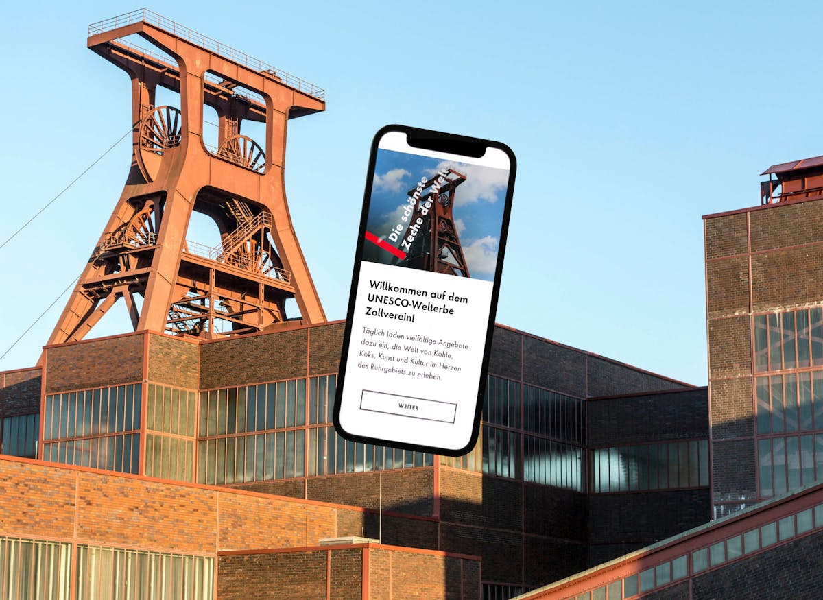 Das Bild zeigt das bekannte Doppelbock-Fördergerüst der Zeche Zollverein in Essen bei Tageslicht mit klarem Himmel im Hintergrund. Im Vordergrund ist ein überdimensionales Smartphone eingeblendet, auf dessen Bildschirm die mobile Website des UNESCO-Welterbes Zollverein geöffnet ist. Auf dem Display ist ein Willkommensgruß zu lesen, der Besucher zur Entdeckung der Welt von Kohle, Koks, Kunst und Kultur im Ruhrgebiet einlädt. Die Gestaltung verbindet historische Industriekultur mit moderner digitaler Information.