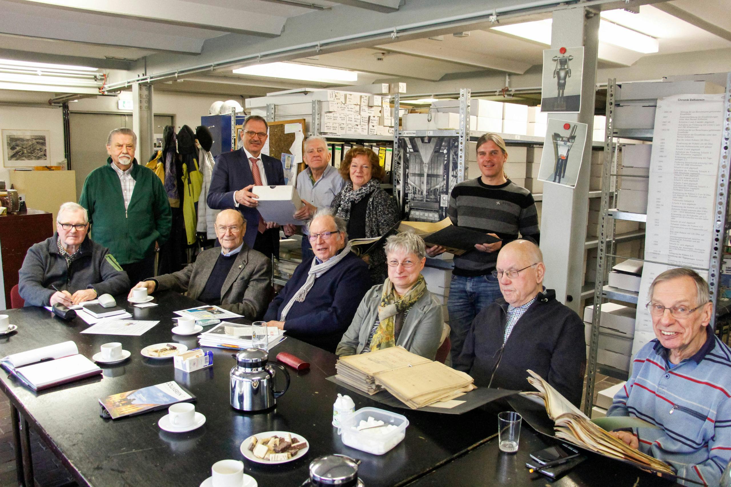 Eine Gruppe von etwa fünfzehn Personen unterschiedlichen Alters sitzt und steht in einem Archivraum um einen großen Tisch. Auf dem Tisch liegen historische Dokumente, Bücher, Tassen und Teller mit Gebäck. Einige Personen blättern in Akten oder halten Archivmaterialien in der Hand. Im Hintergrund sind Regale mit beschrifteten Archivkartons und Aufnahmen von Industrieanlagen zu sehen. Die Atmosphäre wirkt gemeinschaftlich und engagiert, als würde die Gruppe an einem historischen Projekt oder einer Dokumentationsarbeit arbeiten.