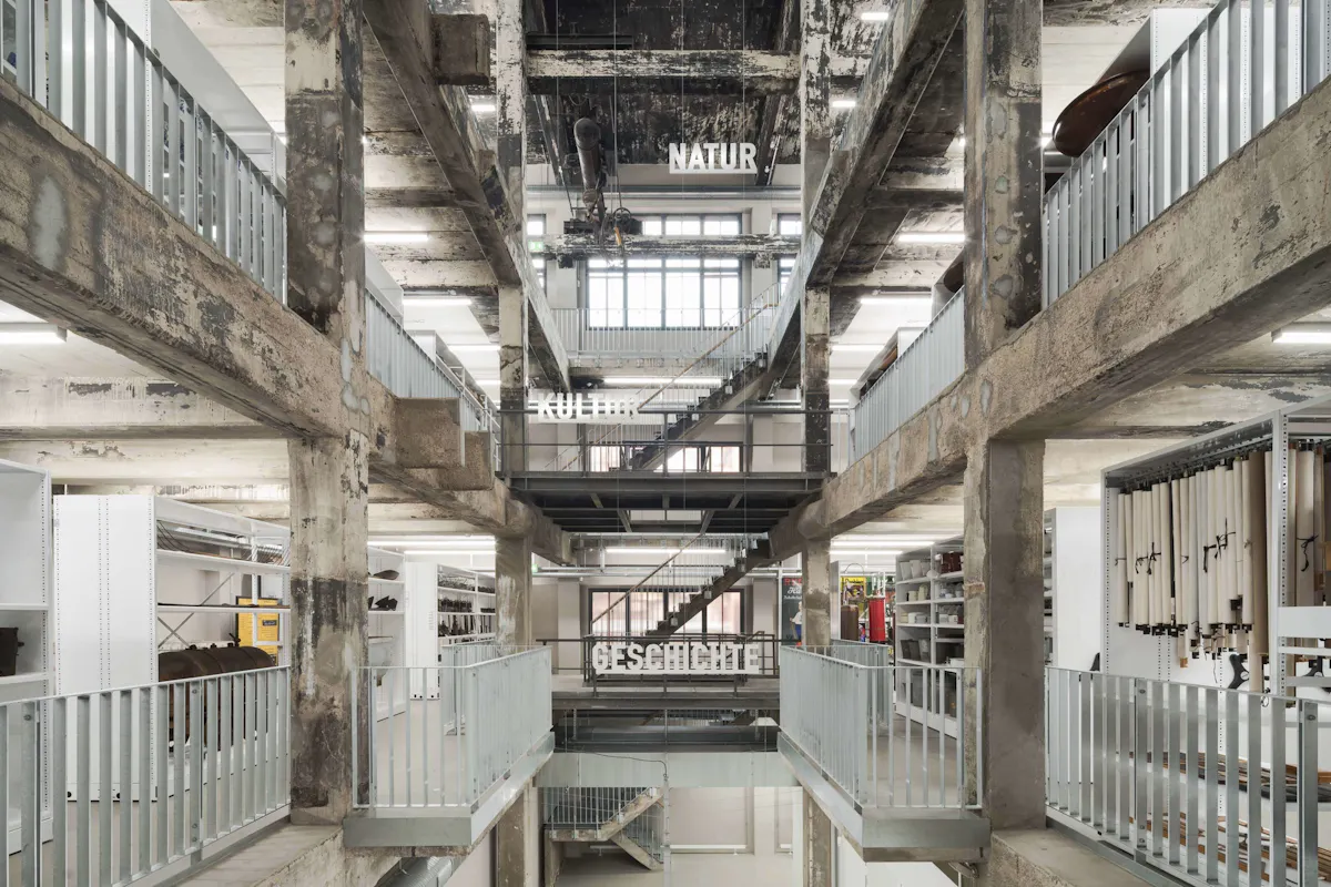 Blick in ein mehrstöckiges Museumsdepot mit industriellem Charakter und sichtbaren Betonstrukturen. Im Zentrum des Bildes befindet sich ein Treppenhaus, das mehrere Ebenen verbindet. An den Geländern sind große weiße Schriftzüge angebracht, die die thematischen Bereiche markieren: unten GESCHICHTE, darüber KULTUR und ganz oben NATUR. Auf jeder Ebene stehen weiße Regale, die mit unterschiedlichen Objekten gefüllt sind – von hängenden Kartenrollen über technische Geräte bis hin zu historischen Gegenständen. Die Architektur wirkt roh und ursprünglich, die Decke ist stark gealtert und weist dunkle Verfärbungen auf. Das Tageslicht fällt durch große Fenster ein und wird durch moderne Deckenleuchten ergänzt.