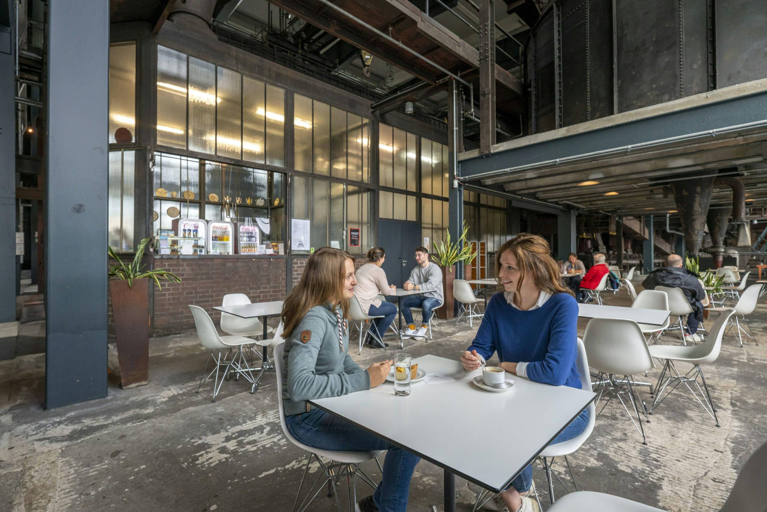 Zwei Frauen sitzen an einem weißen Tisch in einem industriell gestalteten Café mit hohen Decken, Metallkonstruktionen und großen Maschinen im Hintergrund. Sie unterhalten sich bei Kaffee und Wasser, umgeben von weiteren Gästen an modernen weißen Stühlen und Tischen. Im Hintergrund ist ein Verkaufstresen mit Getränken und Snacks sowie ein paar Pflanzen in hohen Pflanzgefäßen zu sehen. Die Atmosphäre ist offen, urban und entspannt.