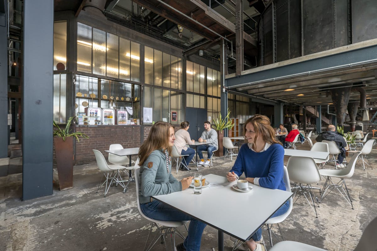 Zwei Frauen sitzen an einem weißen Tisch in einem industriell gestalteten Café mit hohen Decken, Metallkonstruktionen und großen Maschinen im Hintergrund. Sie unterhalten sich bei Kaffee und Wasser, umgeben von weiteren Gästen an modernen weißen Stühlen und Tischen. Im Hintergrund ist ein Verkaufstresen mit Getränken und Snacks sowie ein paar Pflanzen in hohen Pflanzgefäßen zu sehen. Die Atmosphäre ist offen, urban und entspannt.