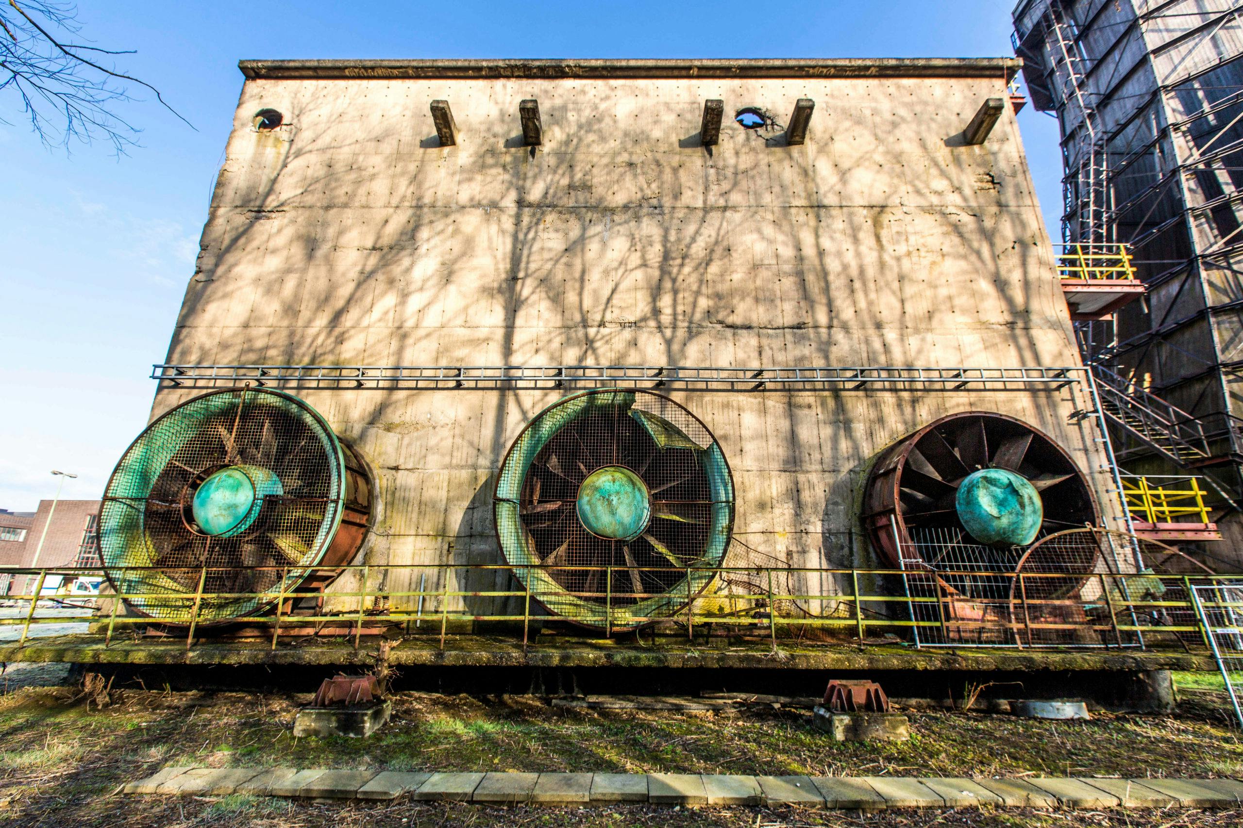 Außenansicht eines alten Industriegebäudes mit drei großen, verwitterten Lüftern oder Ventilatoren in rostigen Metallgehäusen, die horizontal nebeneinander an der Betonfassade montiert sind. Die Lüfter sind mit Drahtgittern gesichert und zeigen deutliche Spuren von Alterung und Korrosion. Das Gebäude wirkt massiv und funktional, mit roher Betonoberfläche und vereinzelten Metallkonstruktionen. Im Hintergrund ist ein weiteres Industriegebäude mit Treppen und Plattformen zu sehen. Die Szene ist bei klarem Tageslicht aufgenommen, wodurch sich die Schatten kahler Bäume deutlich an der Fassade abzeichnen.