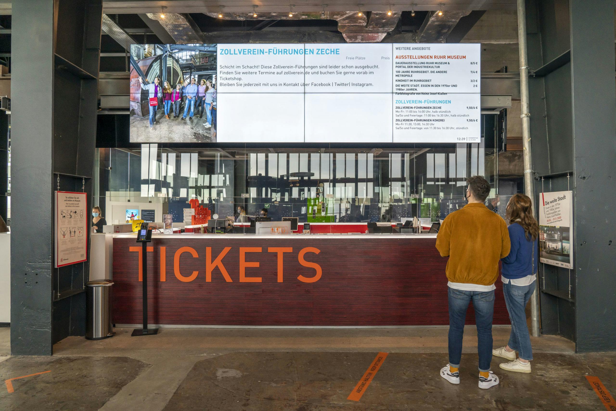 Zwei Personen stehen vor einem modernen Ticketschalter mit der Aufschrift TICKETS in großen orangefarbenen Buchstaben. Über dem Schalter hängt ein großer Bildschirm, der Informationen zu Führungen und Ausstellungen auf dem Gelände der Zeche Zollverein anzeigt. Auf dem Bildschirm sind unter anderem Zollverein-Führungen, Preise und Zeiten zu sehen sowie ein Foto einer Besuchergruppe im Bergwerk. Hinter dem Schalter befinden sich Mitarbeiterinnen und Mitarbeiter in einem hellen, gläsernen Empfangsbereich. Die Umgebung wirkt industriell und modern gestaltet, mit Sichtbeton und offenen Stahlträgern. Links und rechts hängen zusätzliche Infotafeln an den Pfeilern.
